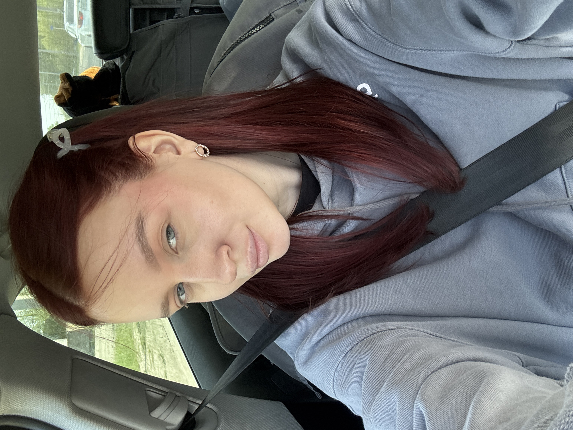A woman with reddish-brown hair is sitting in a car, wearing a light blue hoodie and a seatbelt. She has a small, clear hair clip in her hair and an earring in her left ear. The car interior is visible, including the door panel and what appears to be a car seat or bag in the back. Outside the window, blurred greenery and a building suggest an urban or suburban environment. It is not possible to confirm the location of Balakhna, Russia from the content in the image.