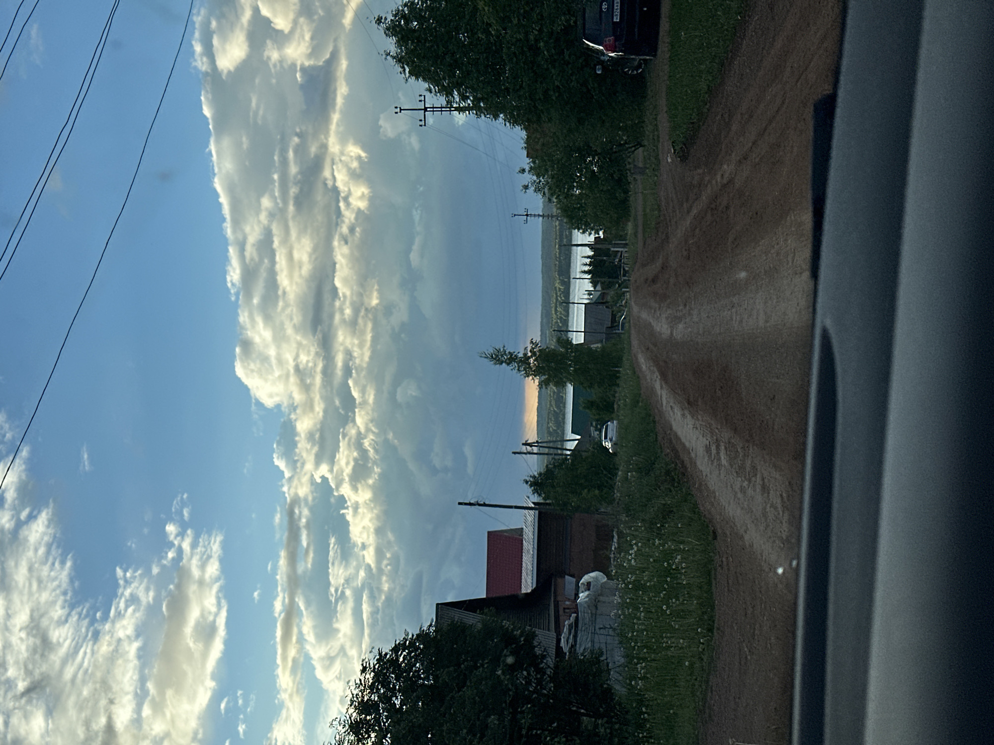 The image shows a dirt road in the city of Ilyinsky, Russia. The road is lined with trees and houses. In the distance, a body of water is visible. The sky is cloudy. A gray vehicle is on the side of the road.