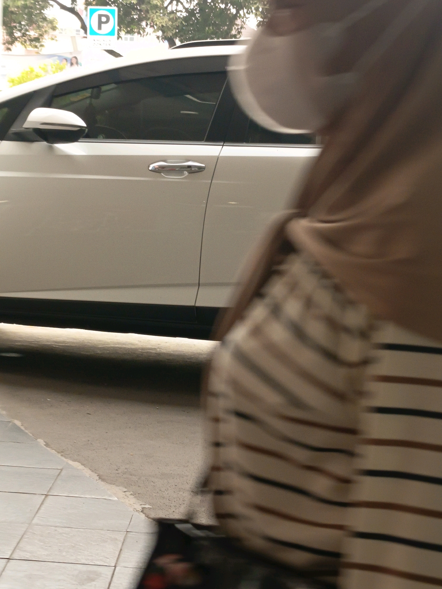 A white car is parked on a street in Tangerang, Indonesia, with a person in a hijab and a striped dress walking past. The car is facing towards the left of the frame, and the person is walking towards the right. The car has a parking sign with a "P" in a blue circle above it. The street is paved with concrete and tiles, and there are trees in the background. The person is wearing a tan hijab and a dress with horizontal stripes in shades of brown and black. The person's face is obscured by a white mask.