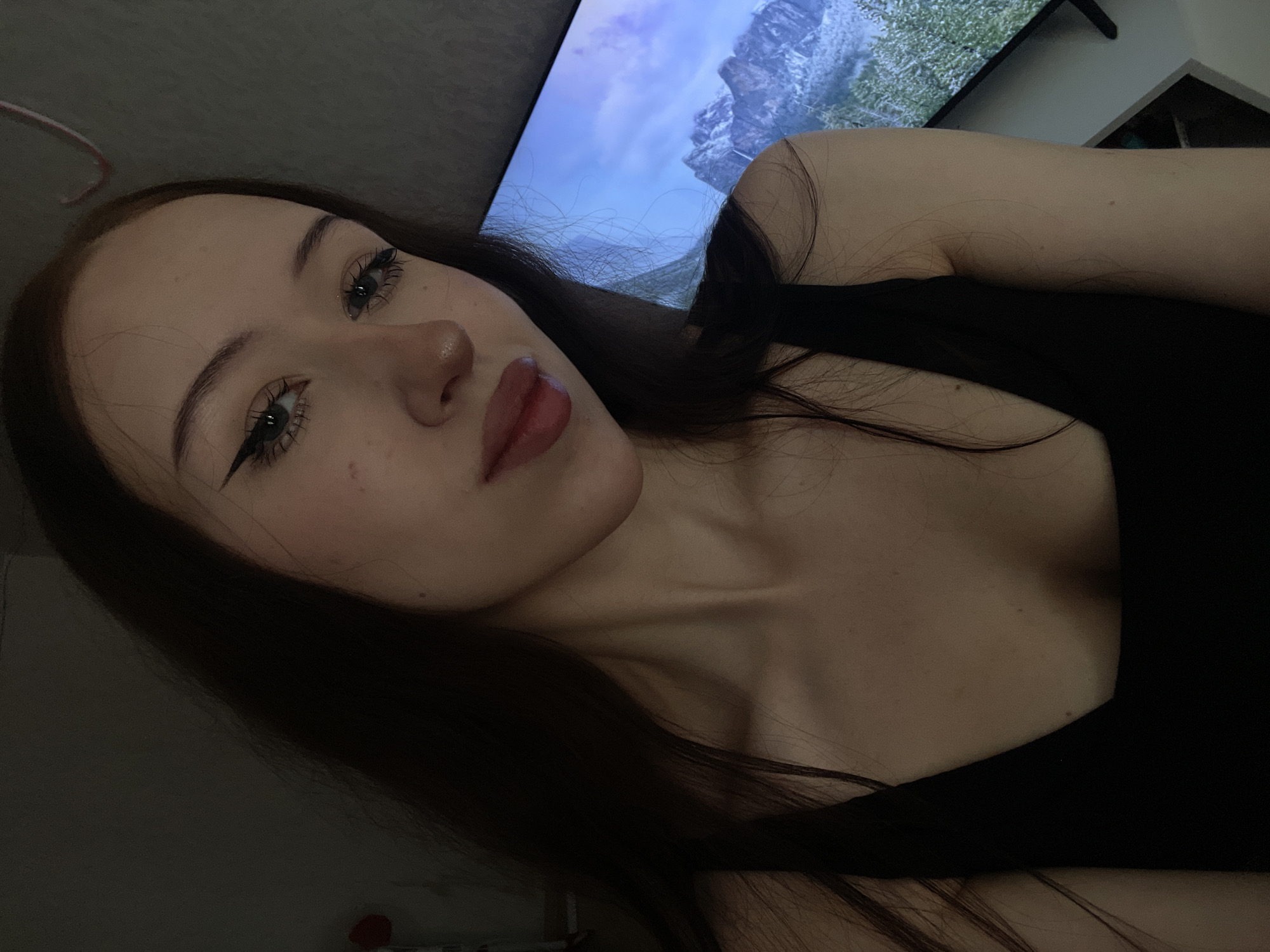A young woman with long, dark hair and pale skin is pictured in a close-up, waist-up shot. She is wearing a black tank top and has winged eyeliner and pink lipstick. Her gaze is directed slightly away from the camera. Behind her, a large screen displays a mountainous landscape with a blue sky. The lighting is dim, with a slight reddish hue on the left side of the image. The scene appears to be indoors, possibly in a residential setting. There is no indication of the specific location within Votkinsk, Russia.