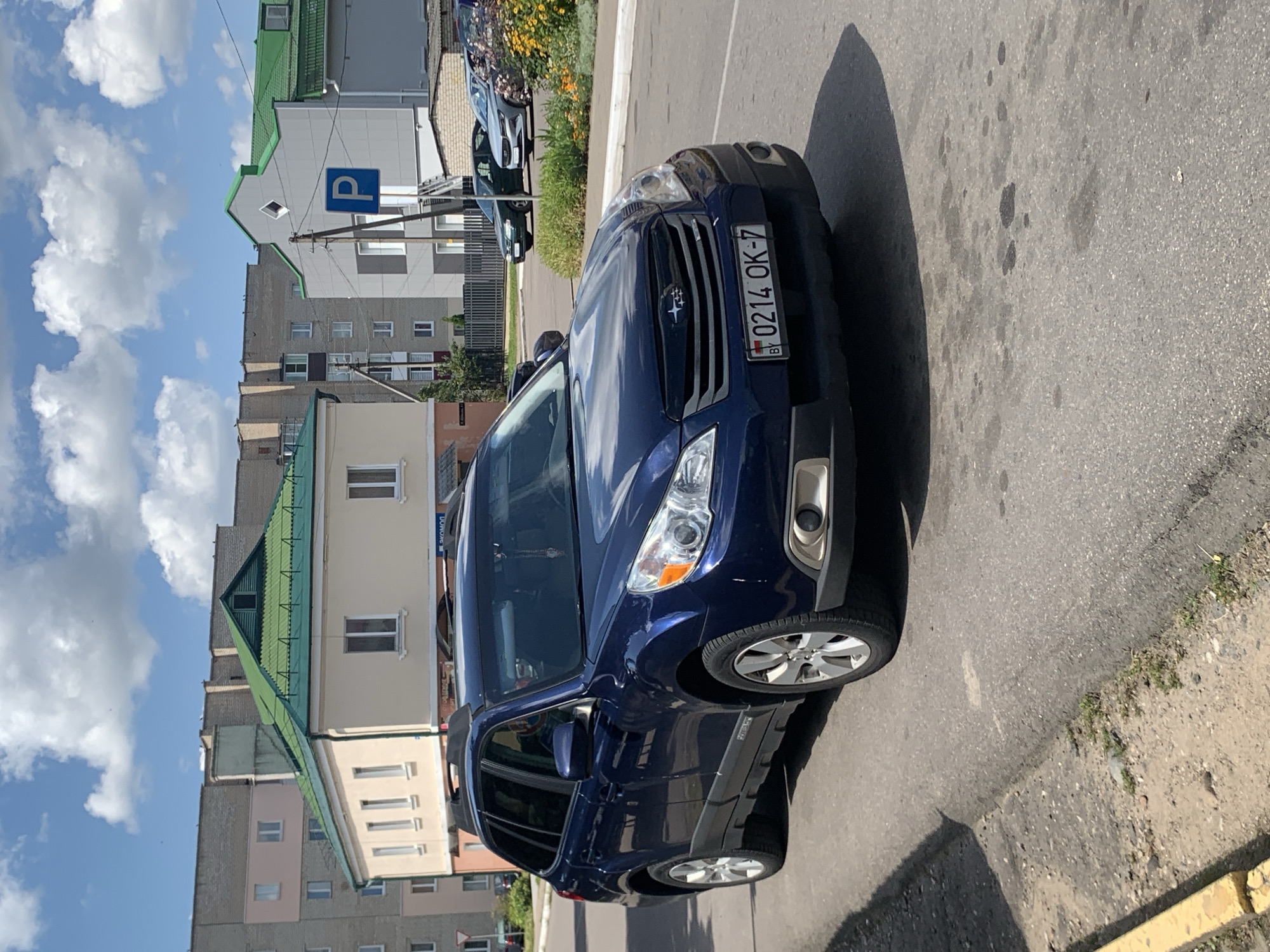 In the city of Kapyĺ, Belarus, a dark blue Subaru Outback station wagon is parked on the right side of a paved road. The car's license plate reads "0214 OK-7" with a "BY" emblem. In the background, there are multi-story buildings with light-colored facades and green roofs. A blue "P" sign indicating parking is visible to the right of the buildings. The sky is blue with scattered white clouds. The road surface shows signs of wear and some dark stains. A yellow painted curb is visible on the left side of the road.