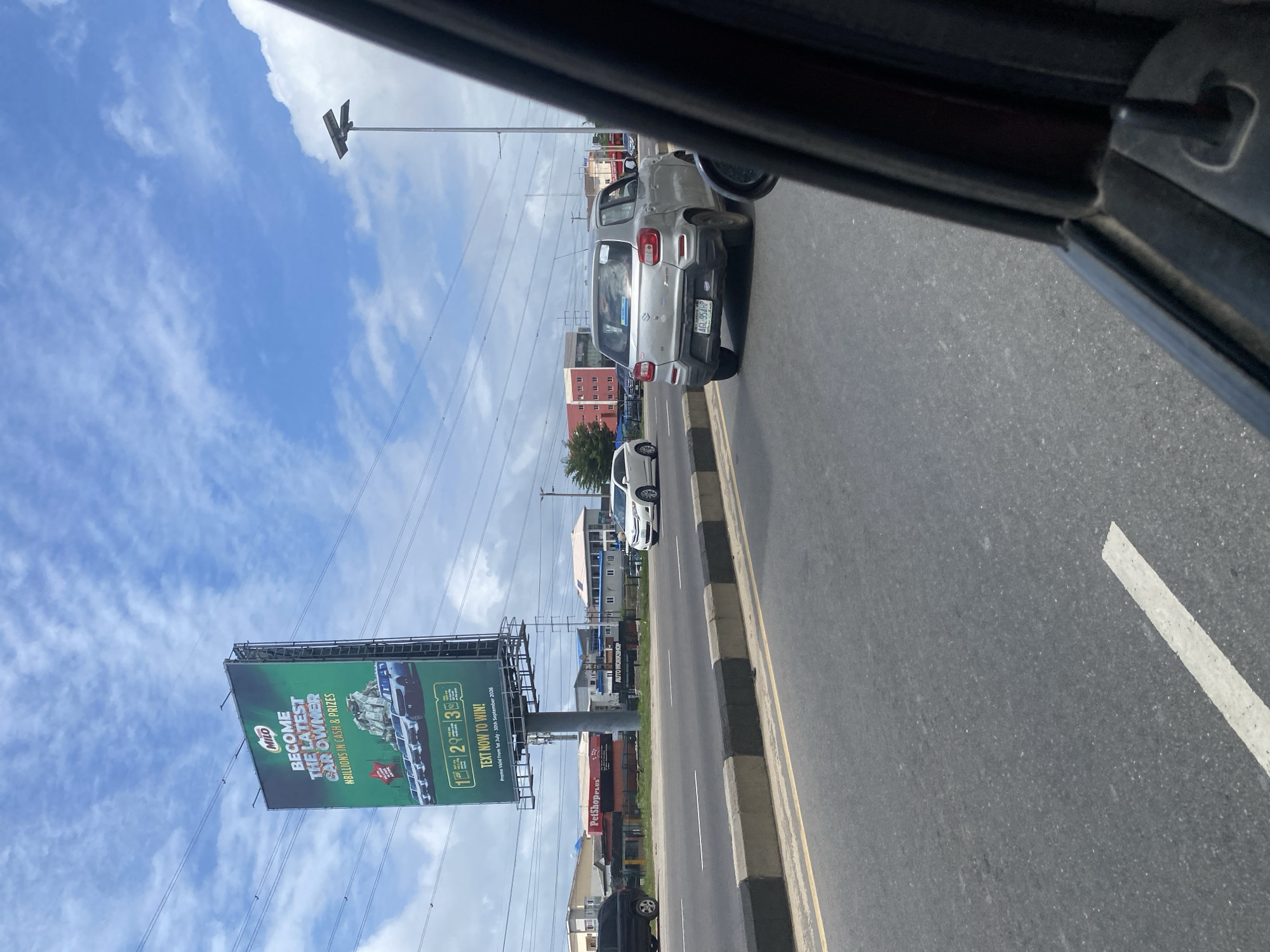 A silver Suzuki S-Presso drives along a paved road on the Lekki Peninsula II, Nigeria, with a billboard for MILO chocolate drink visible in the background. The billboard displays "BECOME THE LATEST CAR OWNER" and depicts stacks of money and cars. Other vehicles and buildings line the roadside, including a "PetShopPlex" and an "AUTO WORKSHOP." The sky is blue with scattered clouds, and overhead power lines are visible. The image is taken from the perspective of a passenger inside another vehicle, with the side mirror and window frame partially in view.