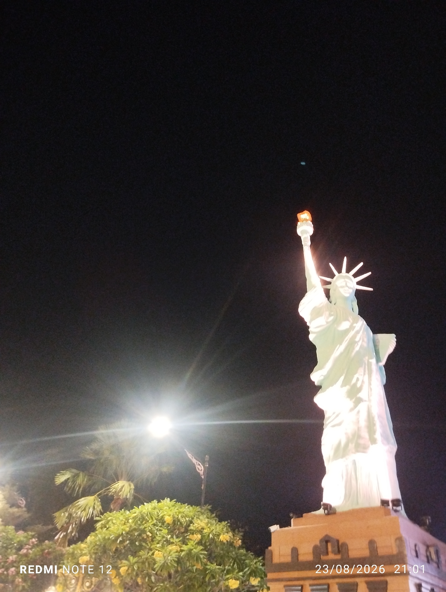 The image, captured at night, shows a white statue of the Statue of Liberty illuminated by lights. The statue is positioned on a brick pedestal. In the foreground, there are trees with yellow flowers and palm trees. Several streetlights are visible, casting bright glows. The bottom left corner of the image displays "REDMI NOTE 12" and the bottom right corner shows the date "23/08/2026" and time "21:01". The scene appears to be an outdoor public space, possibly a park or a landmark in the city of Madiun, Indonesia, given the device information.
