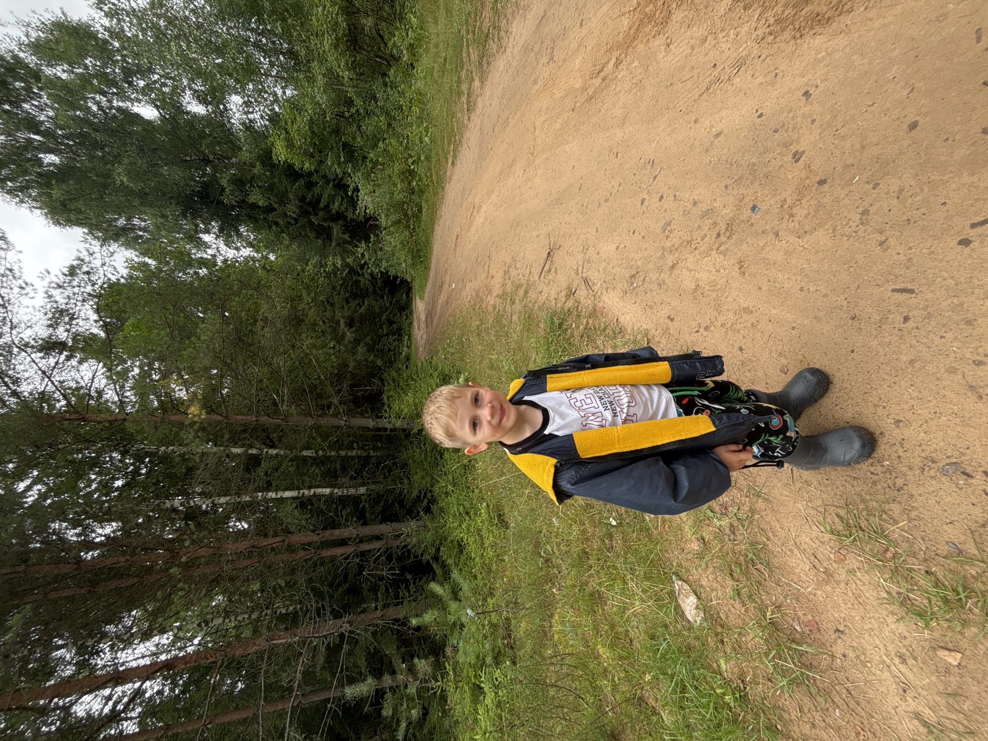 A young boy with blond hair, wearing a dark jacket with a yellow hood, a white t-shirt, patterned pants, and grey rubber boots, stands on a dirt road in a forest. The road curves to the left and disappears into dense green trees on both sides. The ground on the left of the road is covered with grass and some scattered debris. The sky is overcast and appears grey. The location is identified as Anteplevo (Okhotinskoe S/P), Russia.