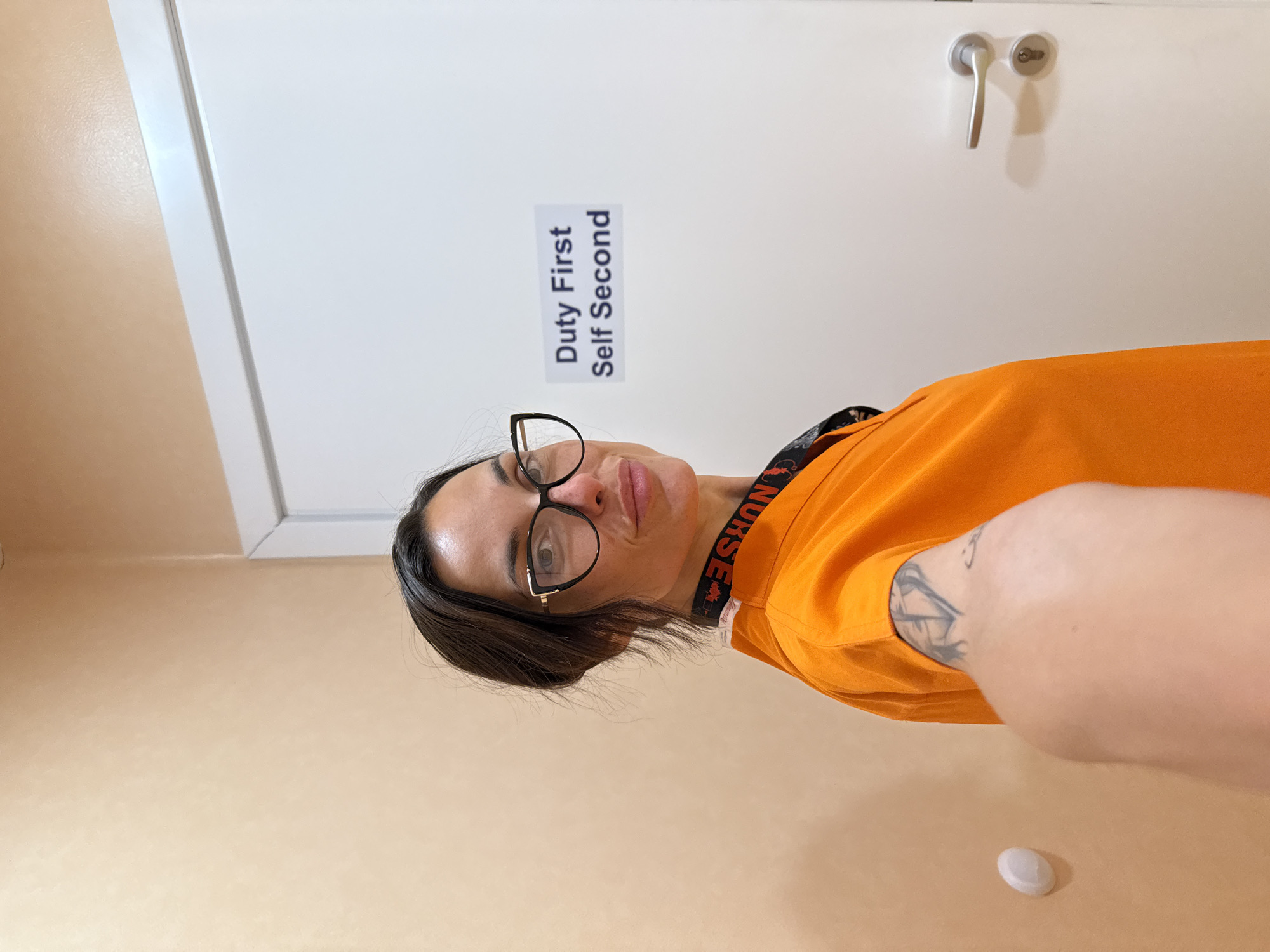 A woman wearing glasses and an orange scrub top is taking a selfie in a hallway. She has dark hair pulled back and is looking at the camera. She is wearing a lanyard that says "NURSE" on it. On the white door behind her, there is a sign that says "Duty First Self Second". There is a door handle on the right side of the door. The walls are a light peach color. A small white circular object is affixed to the wall on the left. A tattoo is visible on her arm. The image appears to be taken indoors, possibly in a hospital or medical facility in Pleven, Bulgaria.