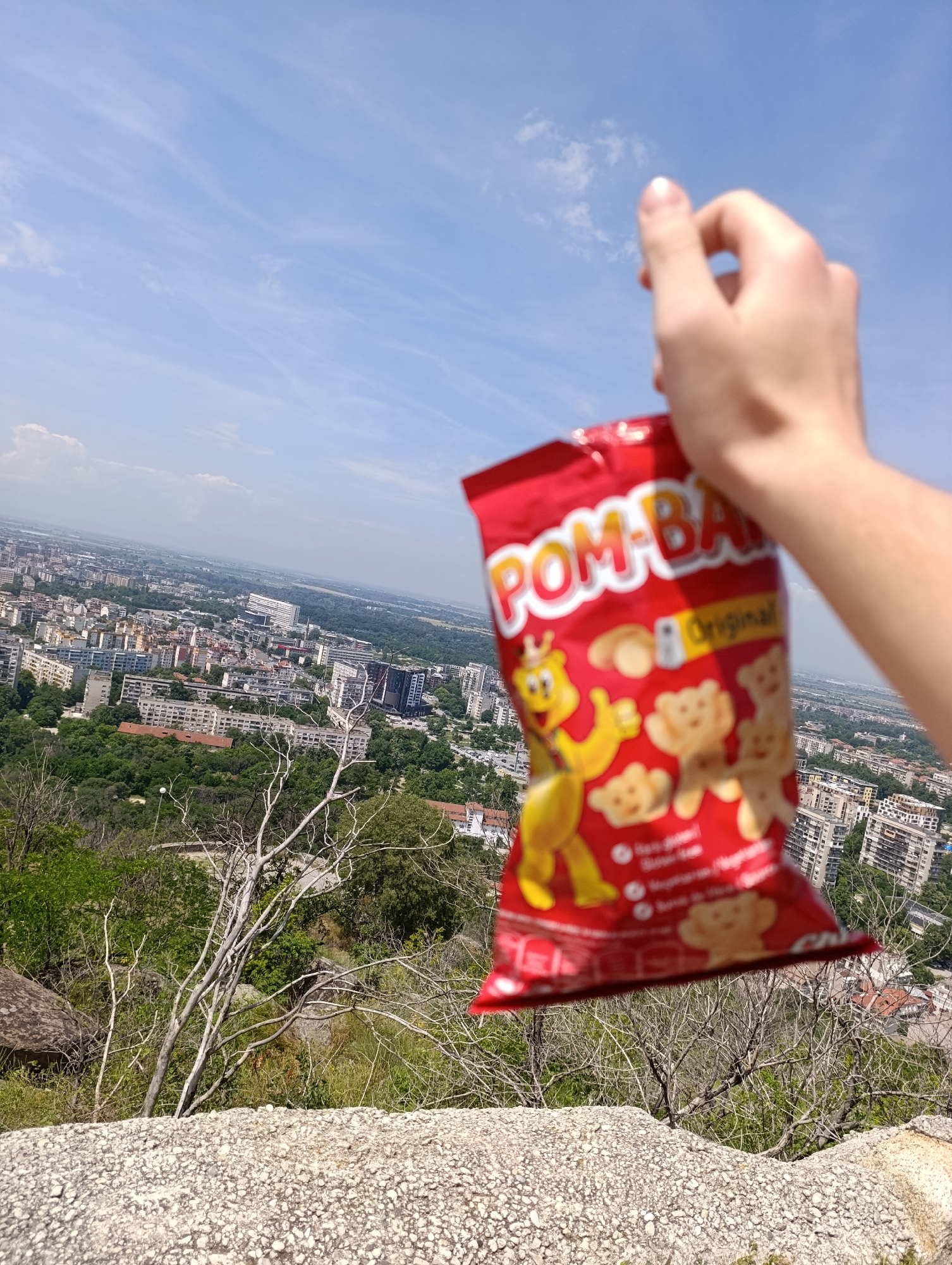 A person's right hand holds up a red bag of "POM-BAR Original" snacks against a clear sky and expansive city view. The hand is light-skinned, and the fingers are slightly bent, supporting the bag.
The snack bag is predominantly red, featuring the brand name "POM-BAR" in large, white, and yellow letters. A yellow bear mascot, giving a thumbs-up, is prominent on the left side of the bag. Several illustrations of light brown, bear-shaped puffed snacks are also visible, along with the word "Original" in a yellow banner. Below the main text, there is some smaller, blurry text, likely indicating ingredients or nutritional information.
The scene is set outdoors, from a high vantage point overlooking a sprawling urban landscape, identified as Plovdiv, Bulgaria. Numerous multi-story residential buildings, interspersed with green parks and trees, stretch across the mid-ground, extending towards a distant, hazy horizon. Roads and other infrastructure are visible within the cityscape. In the immediate foreground, a light-colored, rocky surface dominates the bottom part of the image, with some dry, sparse tree branches and green foliage growing from the rocky terrain.
The time of day is bright, likely mid-day or early afternoon, with clear sunlight illuminating the scene. The sky is a light blue with scattered, thin white clouds. The overall mood suggests a pleasant outdoor break or a moment of appreciation for the view and a snack. The activity depicted is simply holding up the snack bag, possibly before or after consuming its contents, while enjoying the panoramic view.