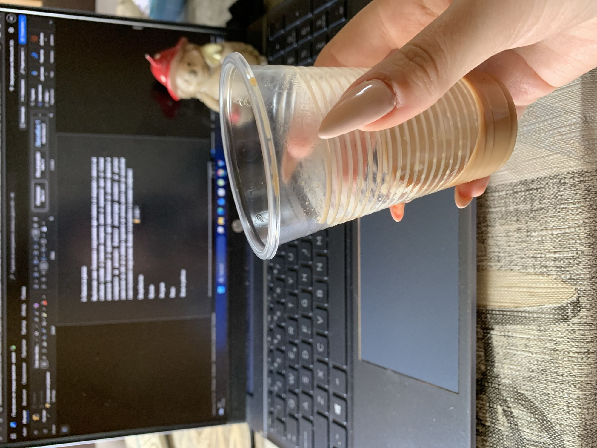 The image shows a person holding a nearly empty clear plastic cup containing a light brown liquid, possibly coffee, in front of a laptop. The laptop screen displays lines of code or text in an editor, with a toolbar visible on the left side. A small, ornamental figurine with a red cap is positioned behind the laptop screen. The laptop is placed on a textured surface, which may be a desk or table. The person's hand is visible, with nails painted in a light beige color. The scene does not provide sufficient information to determine the location of Lehchevo, Bulgaria.