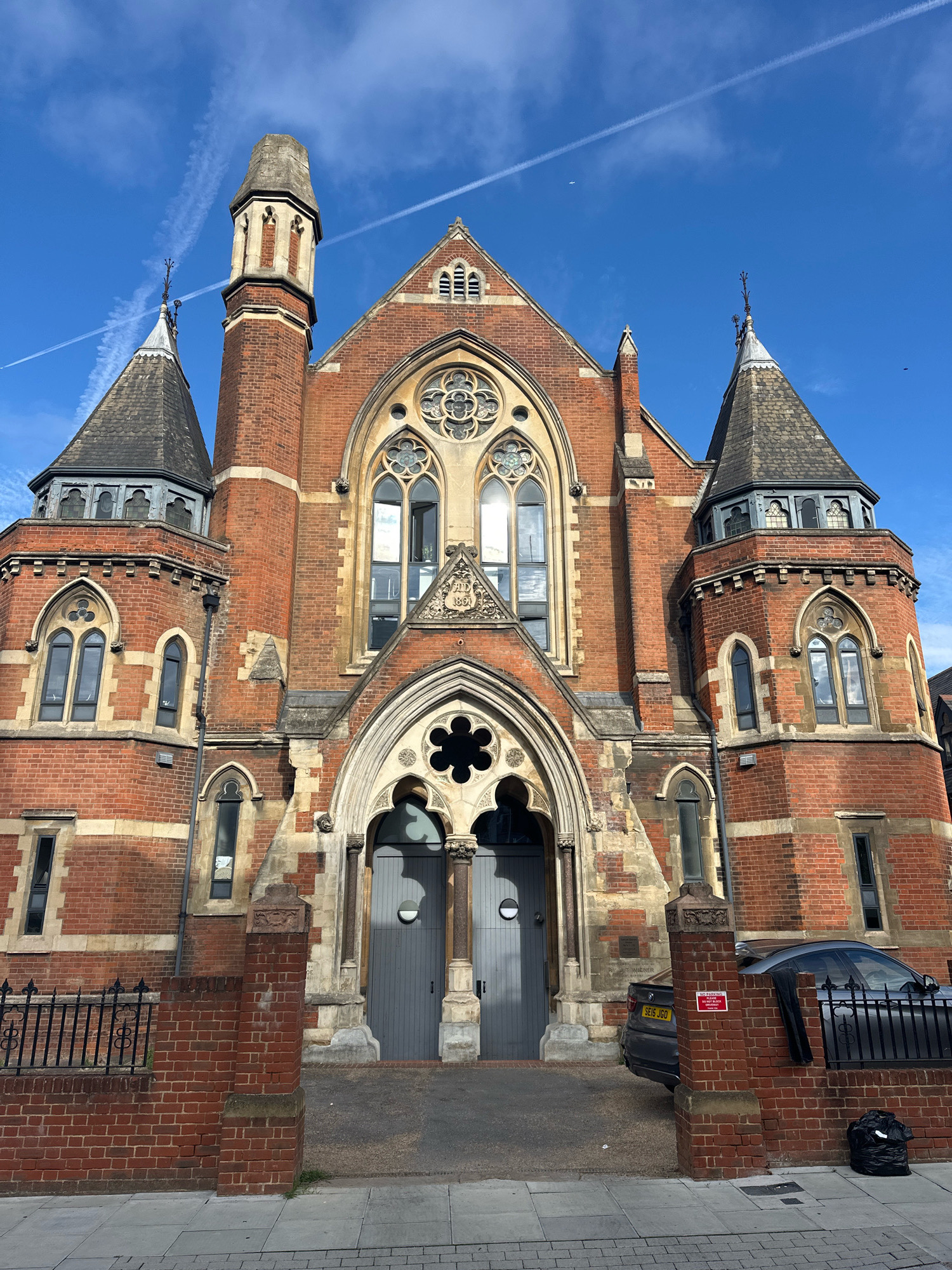 A brick building with Gothic architectural features is pictured on a sunny day in London, United Kingdom. The building has two conical towers with spires and a central section with pointed arches and decorative windows. The entrance features double grey doors. A black metal fence runs along the front of the property, and a grey car is parked behind it. A black trash bag sits on the sidewalk in the foreground. The sky is blue with white contrails from an airplane.