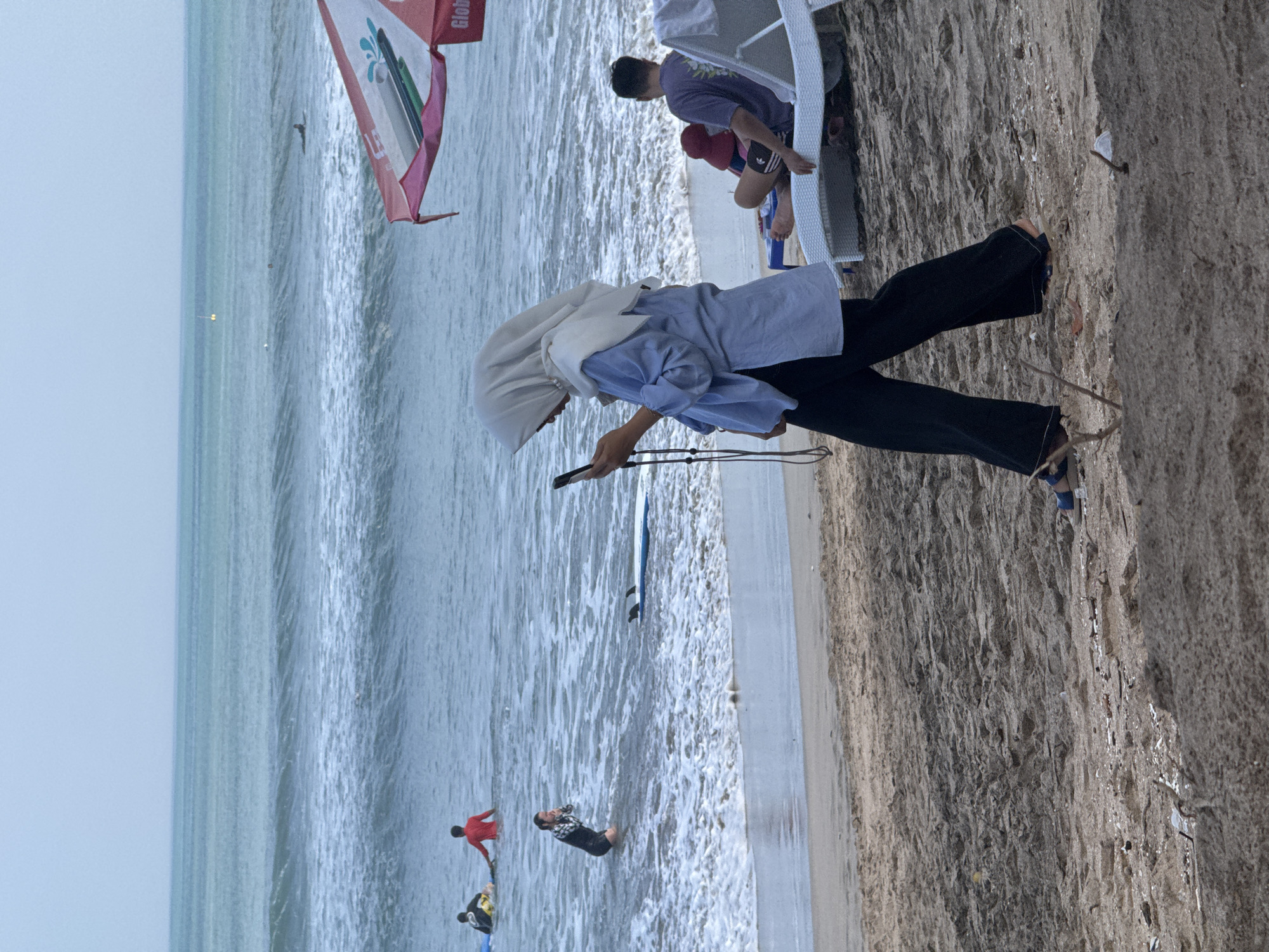The image depicts a beach scene in Kuta, Bali, Indonesia. In the foreground, a woman wearing a light blue long-sleeved top, black wide-leg pants, and a light-colored hijab is walking along the sandy beach while looking at her smartphone. She is also wearing blue flip-flops. Behind her, on a white lounge chair, a person wearing a purple t-shirt and dark shorts is seated, facing away from the camera. A red hat is visible on another person sitting nearby. Further back, in the shallow water, a person in a black wetsuit is kneeling, and a blue surfboard with fins is floating in the water. Other surfers can be seen in the distance on surfboards in the ocean. The ocean has moderate waves, and the sky is overcast. An umbrella with the word "Global" visible on it is partially in the upper right corner of the frame.