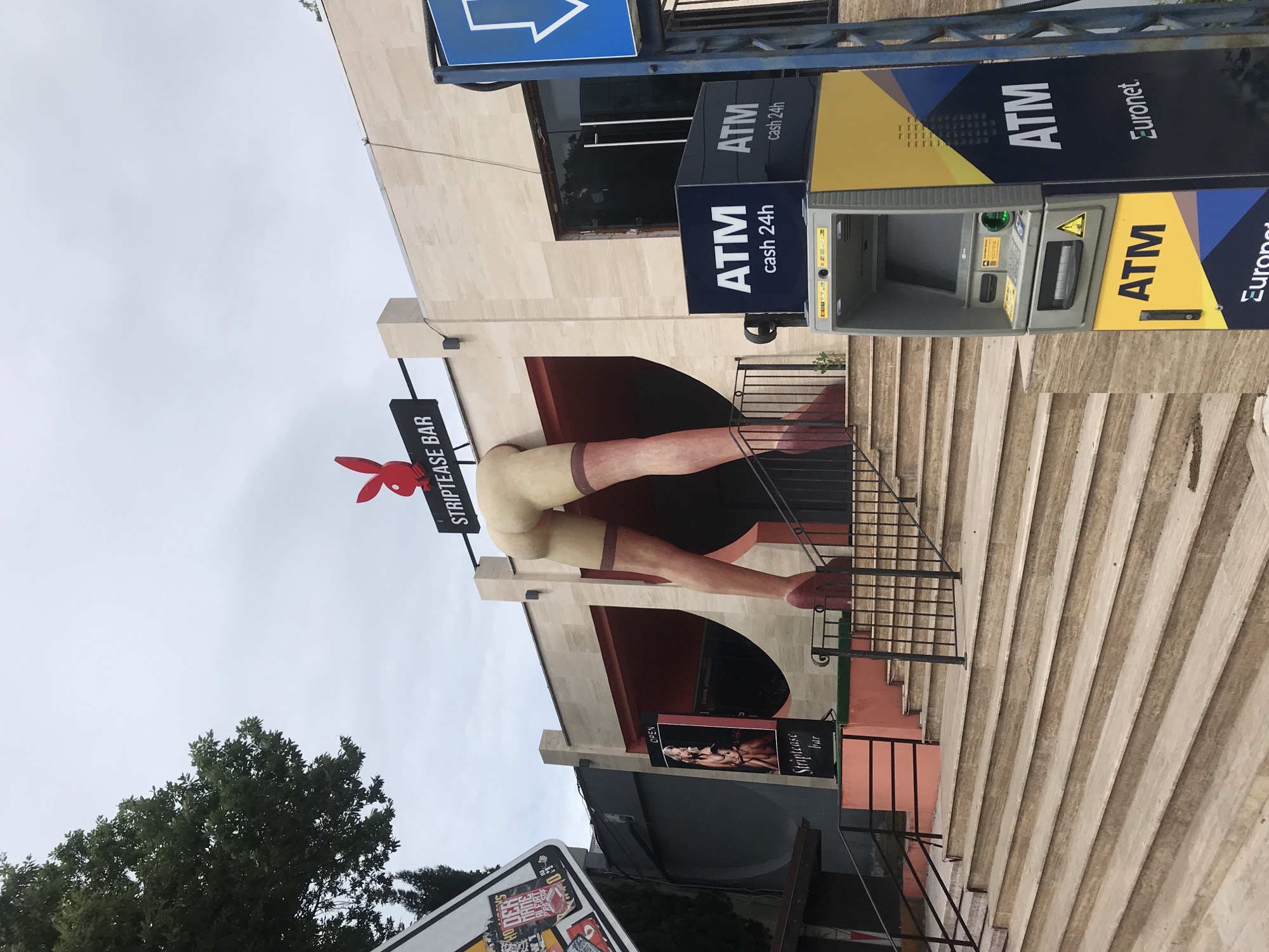 An outdoor scene depicts the exterior of a "Striptease Bar" in Varna, Bulgaria. The bar's facade features a large, stylized sculpture of a woman's legs wearing stockings and high heels, emerging from an opening. Above the entrance, a sign reads "STRIPTEASE BAR" and is adorned with a red Playboy bunny logo. To the left of the entrance, a vertical sign displays the text "Striptease bar" and an image of two women. Several steps lead up to the bar's entrance. To the right of the steps, an ATM machine is visible, with branding for "ATM cash 24h" and "Euronet." The sky is overcast, and a large tree is present on the left side of the image.