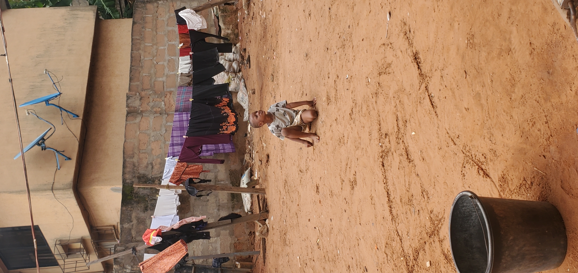 A young child is crouching on the ground of an unpaved yard in Onitsha, Nigeria. Clothes are hanging on a makeshift line, which is strung up in front of a building with satellite dishes. A dark-colored bucket is near the right edge of the scene.
