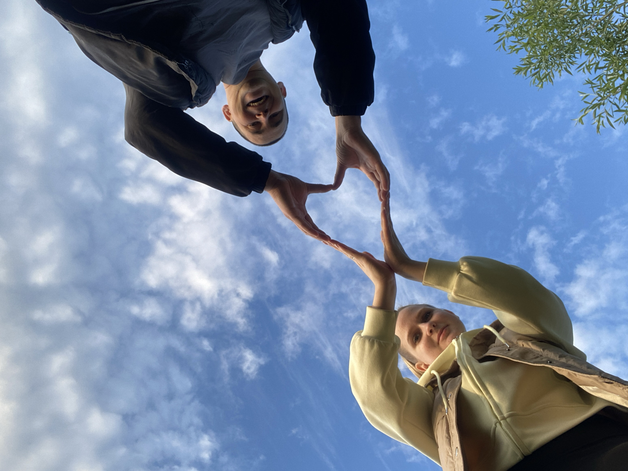 Two individuals are positioned in the frame, forming a heart shape with their hands against a backdrop of a blue sky with scattered white clouds. The perspective is from below, looking upwards.
The first individual, positioned at the top of the frame, is a light-skinned male with short hair, a dark jacket, and a grey shirt. He is smiling. His hands form the upper portion of the heart shape.
The second individual, at the bottom of the frame, is a light-skinned female wearing a light yellow hooded sweatshirt and a dark vest. Her hands form the lower portion of the heart shape.
In the upper right corner, there is a glimpse of green leaves, possibly from a tree. The image suggests an outdoor setting in Chaikovsky, Russia, with a bright, daytime atmosphere.