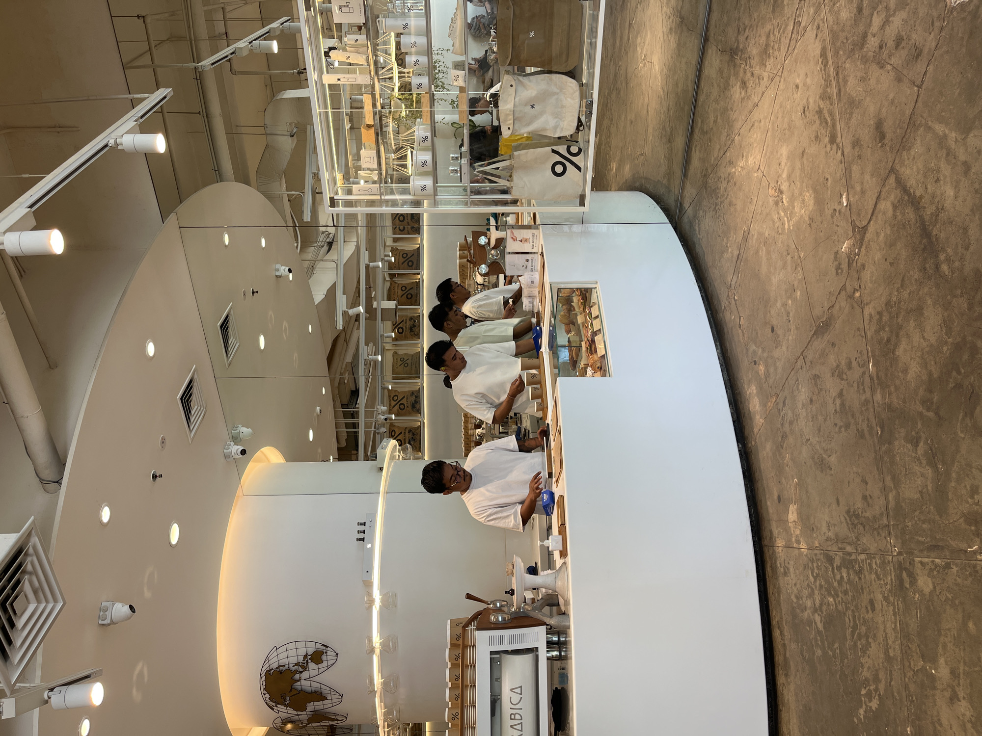 The image depicts the interior of a modern coffee shop, likely % Arabica, located in Kuta, Indonesia. Three young men, dressed in white t-shirts, are visible behind a white curved counter. The man on the left, wearing glasses, is interacting with a customer off-camera, possibly taking an order or preparing a drink. The other two men appear to be assisting with tasks behind the counter.
The counter is well-lit and features a coffee machine labeled "ARABICA" on the left side. A display case filled with pastries and baked goods is visible in the foreground. Behind the counter, shelves are stocked with various merchandise, including tote bags with the distinctive "%" logo, coffee cups, and other branded items. The ceiling has exposed industrial elements, including pipes and ventilation systems, along with track lighting. Security cameras are mounted on the ceiling. The floor is a polished concrete surface, showing signs of wear and some cracks. In the background, a large display with a world map graphic is mounted on a curved white pillar. The overall ambiance is clean, minimalist, and contemporary, consistent with a high-end coffee chain.