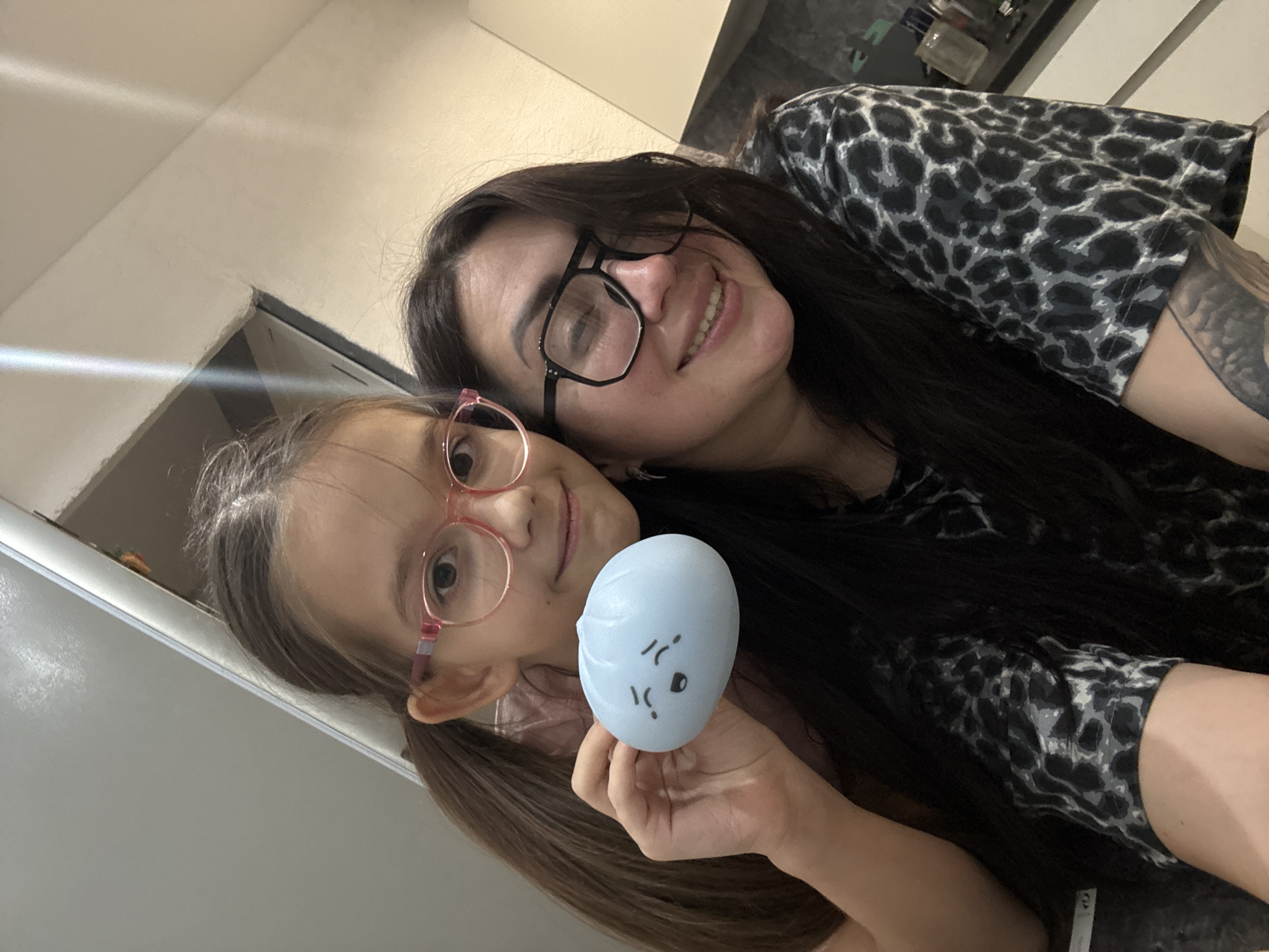 An indoor, tilted, medium-close-up photo shows two females, an adult woman and a young girl, smiling at the camera.
The adult woman, positioned on the right and slightly behind the girl, has long dark hair, black-framed glasses, and is wearing a black and grey leopard print top. A tattoo is visible on her left arm. She is smiling broadly.
The young girl, in the foreground on the left, has light brown hair tied in pigtails and wears pink-framed glasses. She is holding up a small, light blue, oval or dumpling-shaped object with a simple cartoon face featuring closed eyes and a downturned mouth. The girl is also smiling.
The background appears to be an indoor setting with light-colored walls and possibly shelves or cabinets, suggesting a domestic environment such as a kitchen or living area. Both individuals convey happy and relaxed emotions. There is no discernible text visible in the image.