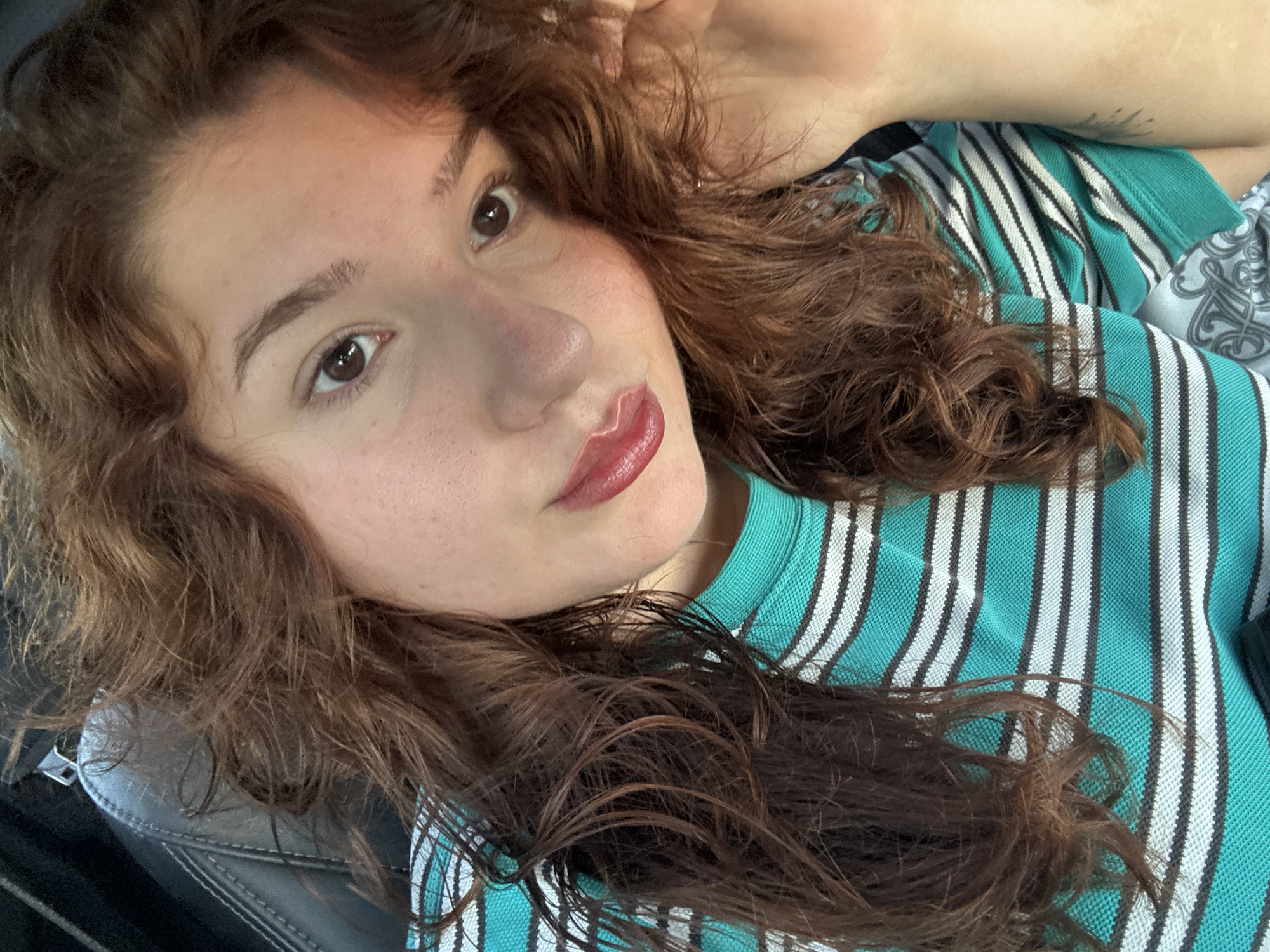 A close-up selfie depicts a young woman with light skin, brown eyes, and reddish-brown curly hair. She is wearing a teal and white striped shirt. Her head is tilted, with her hand resting behind it. She has lipstick on. The location is not discernible as Elhovo, Bulgaria based on visual evidence.