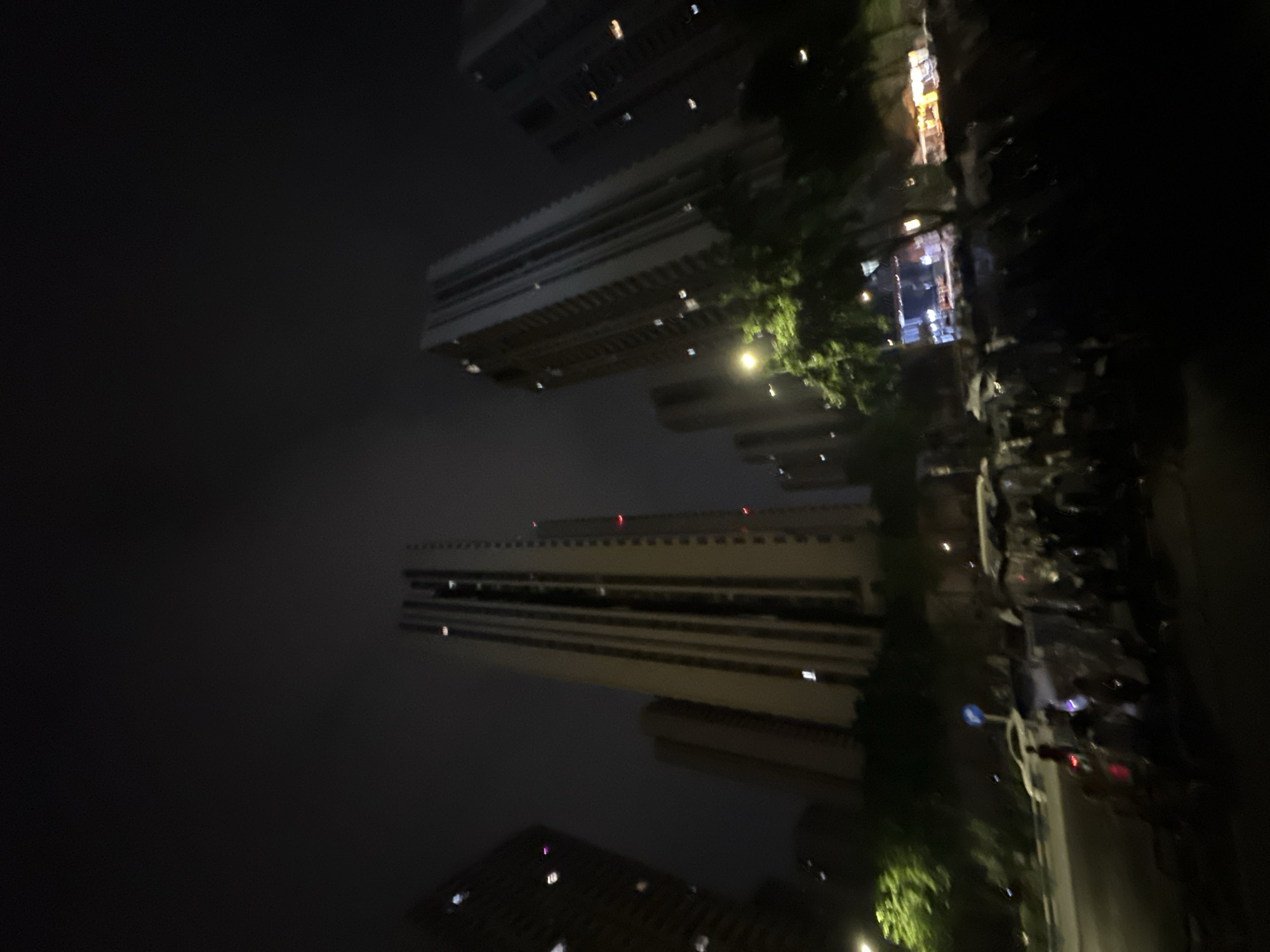 The image depicts a nighttime urban scene in the Chongqing High-tech Zone, China. The sky is dark and overcast, with minimal light penetration. Dominating the upper and middle ground are multiple tall, modern residential or commercial buildings, characterized by their vertical lines and numerous illuminated windows. Some buildings exhibit red lights, likely aircraft warning beacons, on their upper sections.
At street level, a roadway is visible, flanked by trees with green foliage illuminated by a bright, yellowish street lamp on the left. A white sedan or SUV is discernible on the road, near a faint blue circular road sign. The right side of the street features a dense concentration of parked two-wheeled vehicles, likely electric scooters or motorcycles, many covered with translucent plastic tarpaulins. Several indistinct human figures are present on the street, some in proximity to the parked vehicles. In the far background, areas of brighter illumination suggest active storefronts or commercial establishments. The overall image quality is characterized by blur, particularly in the foreground and areas of motion.