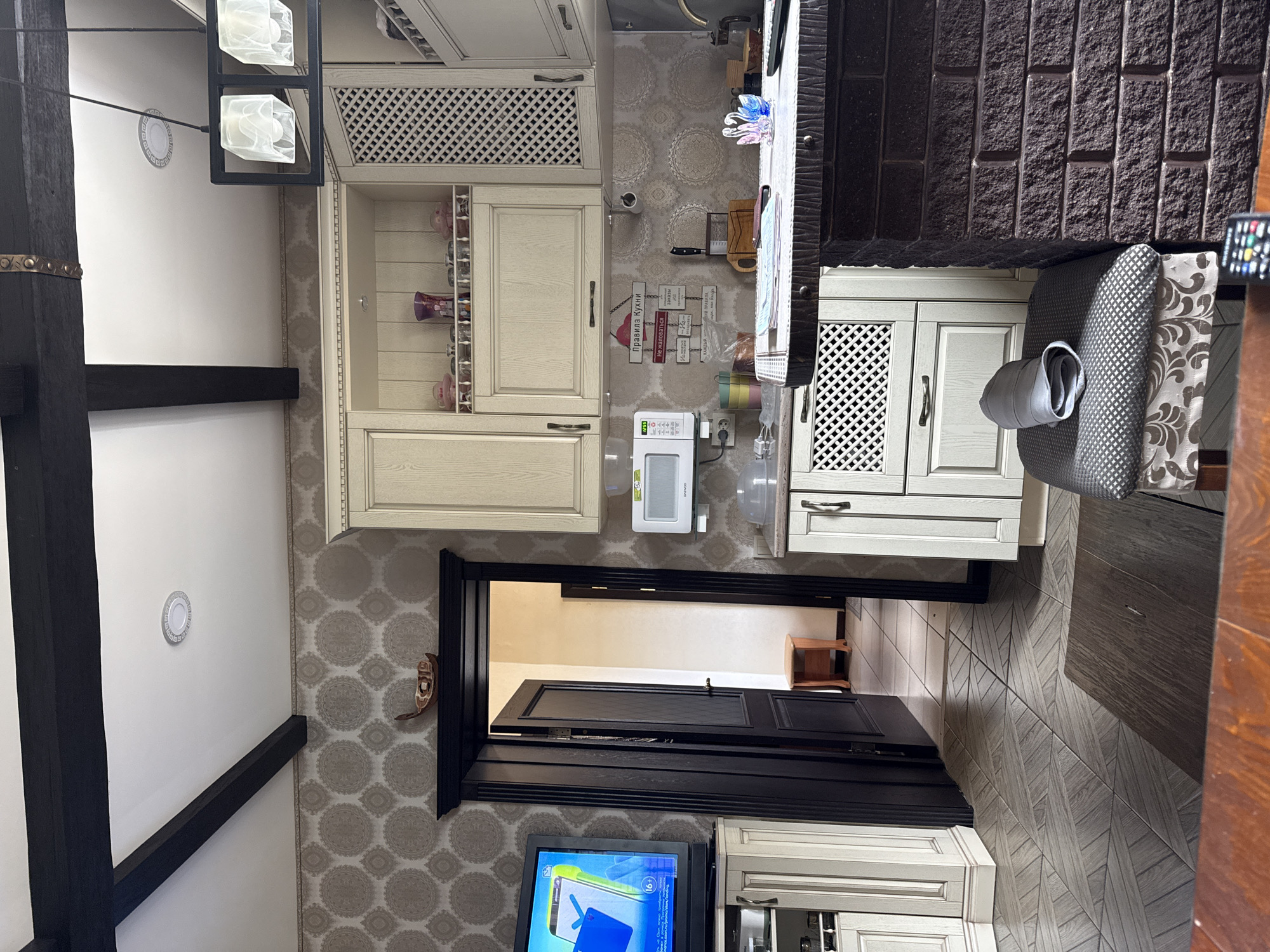 This media file captures an indoor scene, likely a kitchen or dining area within a home in Krapivka, Russia. No people are visible.
The room features light-colored, possibly cream or light wood, kitchen cabinetry, some sections of which have decorative lattice-patterned fronts. A white microwave oven, partially branded "DEWOO" or "DAEWOO," sits on a countertop. Above it, a water dispenser is visible. The walls are adorned with light-colored wallpaper featuring a repeating circular/mandala pattern. A collection of decorative wooden signs hangs on one wall, forming "Kitchen Rules." Visible text on these signs includes "ПРАВИЛА КУХНИ" (Kitchen Rules), "Не жаловатся" (Do not complain), "Пришла - убери" (Came - clean up), "Не наедайся" (Don't overeat), and phrases relating to chores like "Мужчина должен" (A man must) and "Посуда - женское дело" (Dishes - women's business).
A dark, possibly brick or stone-clad wall section separates the main kitchen area from a dining space. A chair with patterned gray and white upholstery is pulled up to a dark-colored table or counter. A remote control is visible on the far right of this surface. Dark wooden beams are prominent in the ceiling structure, and a modern light fixture with two square, frosted glass shades hangs down. The floor is tiled with a gray, wood-look pattern laid in a herringbone or similar arrangement. A black-framed doorway leads to another room, where a small wooden stool is visible. Below this opening, a TV screen displays colorful graphics and a possible age rating "16+".