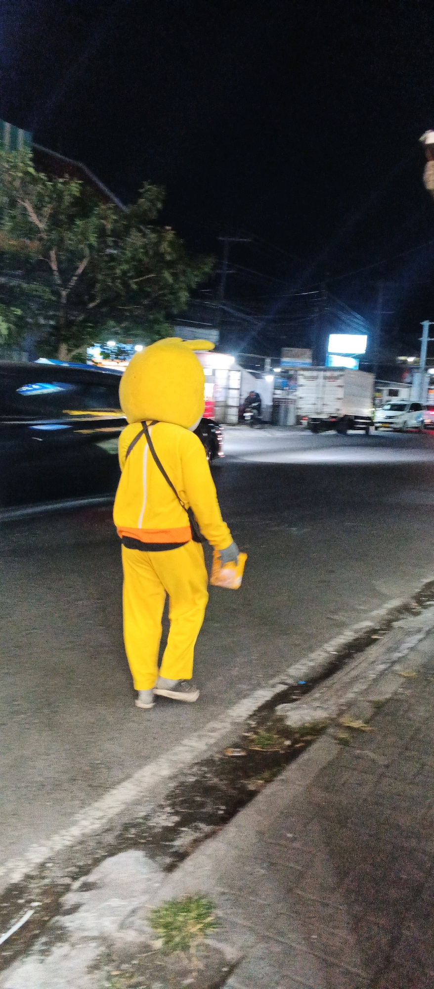 A person dressed in a full-body yellow mascot costume, resembling a stylized duck or chicken, walks on a paved road at night. The costume has a large, rounded head and is paired with yellow pants and a jacket. A black strap crosses the chest of the costume, and the person is holding a small, orange object in their gloved hand. The background suggests an urban or semi-urban setting, with streetlights, trees, buildings, and vehicles visible. Specifically, the location is identified as Karangploso, Indonesia. The scene is captured from a slightly elevated, rear-facing perspective.