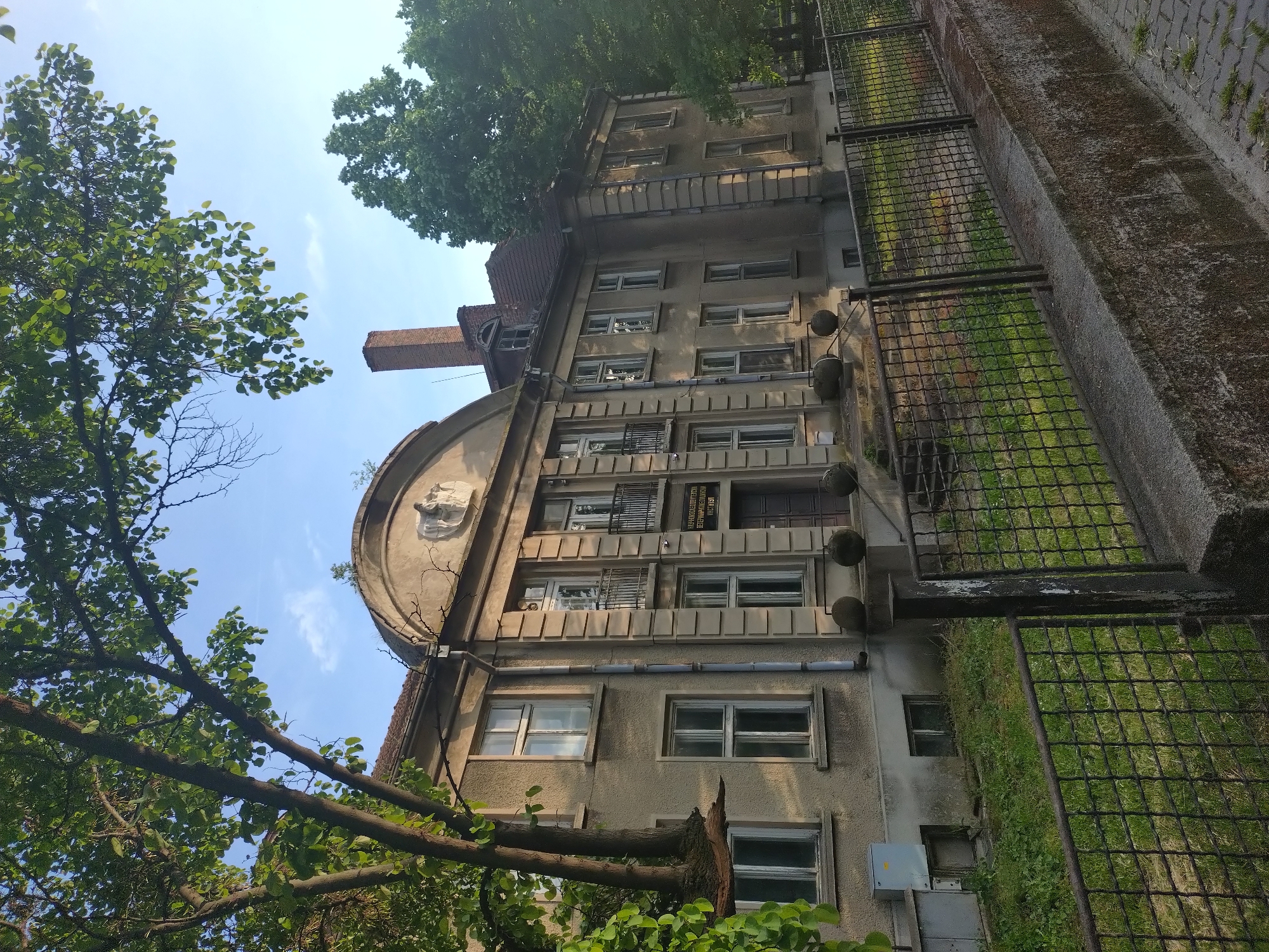 The image shows a multi-story building with a prominent arched facade element topped with a sculpted emblem in Stara Zagora, Bulgaria. The building appears aged, with a rough textured exterior and multiple windows, some with metal grilles. A tall brick chimney is visible on the left side of the building. A metal fence runs along the right side of the building, separating it from a patch of grassy area. Trees frame the building on either side, partially obscuring the view. The sky is visible in the background, ranging from blue to a lighter hue.