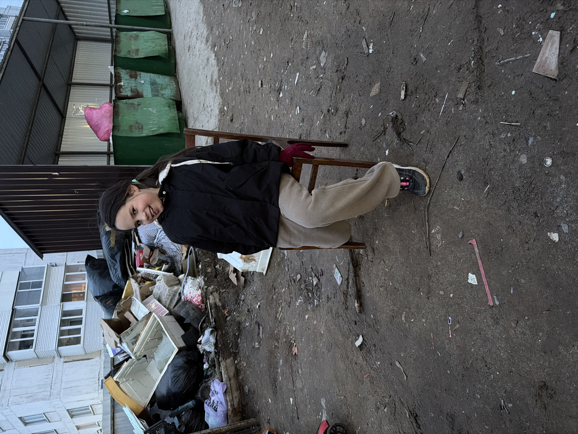 A full-length portrait features a young girl, likely pre-teen, seated on a simple wooden chair in a neglected outdoor environment in Chaikovsky, Russia. She has long dark hair and is wearing a black puffer jacket, light beige sweatpants, and dark sneakers with pink accents. Red gloves are visible on her hands as she rests them on the chair's backrest. The girl is looking directly at the viewer with a noticeable smile on her face.
The setting appears to be a communal waste disposal area. To the left of the girl is a large, overflowing pile of garbage, consisting of black plastic bags, cardboard boxes, broken household items including a white cabinet or drawer unit, and various other refuse. Behind this trash pile, a multi-story residential building with white paneled walls and numerous rectangular windows rises.
The ground around the girl and the trash is a mix of damp, dark earth, mud, and gravel, heavily littered with small pieces of debris, including paper, plastic, and twigs. In the background, beyond the trash pile, are several industrial-looking green metal structures, possibly covers for large dumpsters or part of a recycling enclosure. A pink heart-shaped balloon is incongruously perched on top of one of these green structures, adding a splash of vibrant color to the otherwise bleak scene.
The lighting is soft and diffused, suggesting an overcast day or perhaps early morning/late afternoon. The overall atmosphere is one of stark contrast between the child's cheerful expression and the dilapidated, trash-filled surroundings. No clearly legible text is visible in the image.