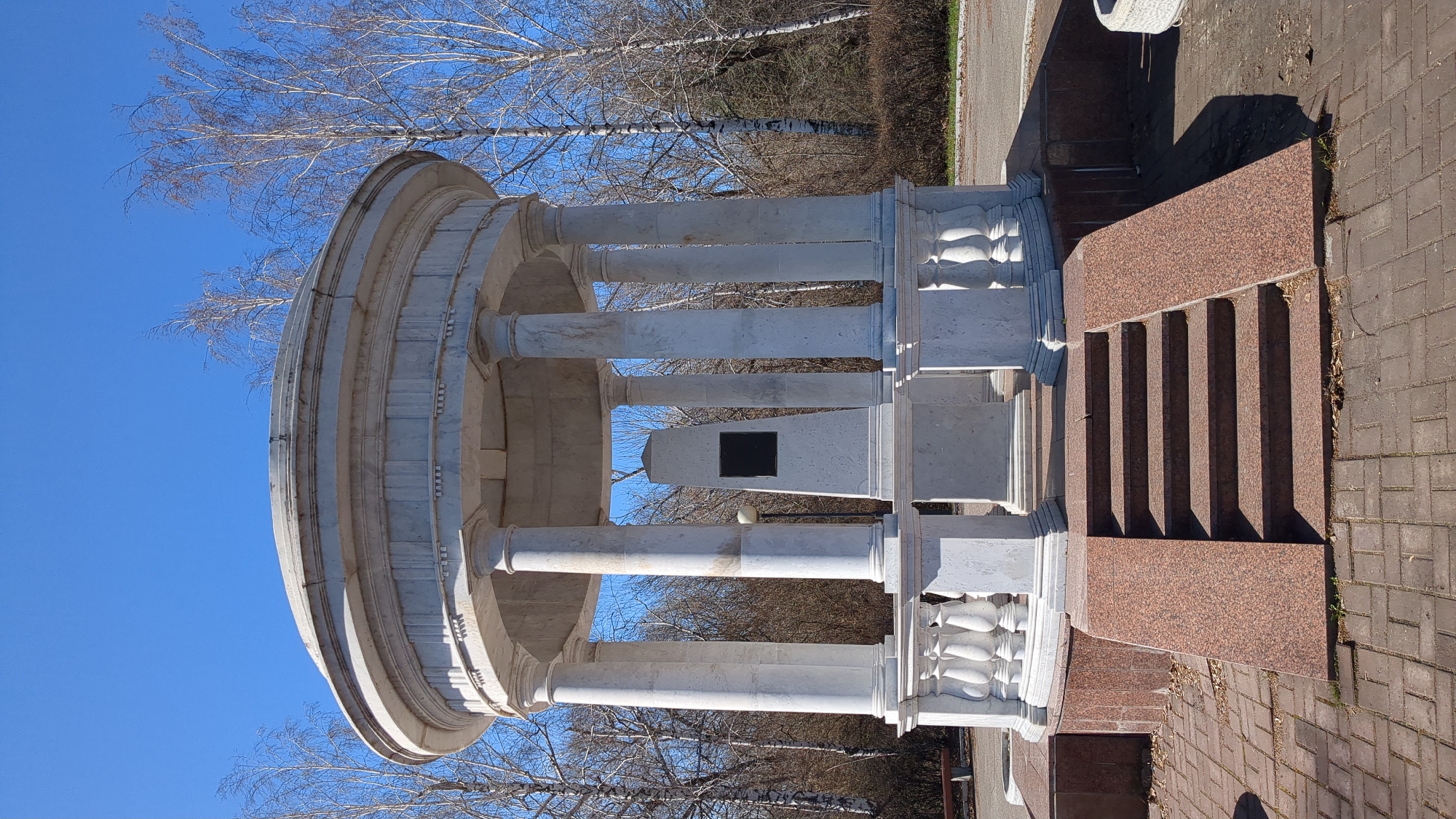 The image depicts a circular white marble gazebo or rotunda-like structure with classical columns, situated on a brown brick-paved area. A small black plaque is visible inside the structure. A staircase with brown granite steps leads up to the structure. Bare trees are visible in the background against a bright blue sky. Based on the structure and location, it is likely the "Gazebo-monument dedicated to the letter 'Yo'" in Ulyanovsk, Russia.