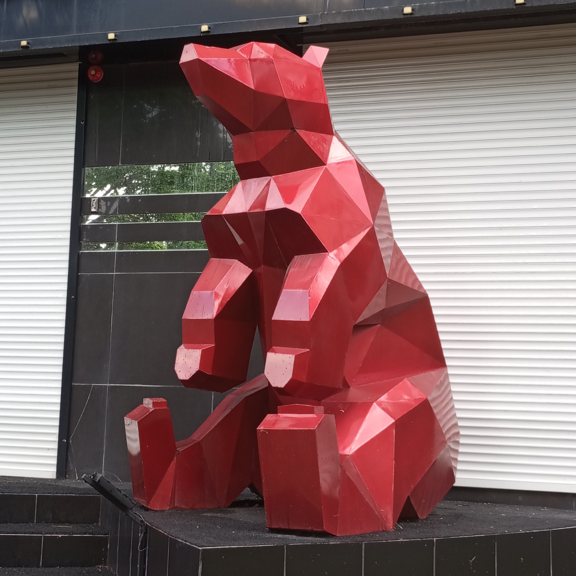 A large, faceted, red bear sculpture sits outside a building. The sculpture is made of geometric shapes and appears to be made of metal. The bear is sitting down with its legs extended in front of it. The building behind the bear has a black facade with white roll-up doors. There is also a reflection of trees and greenery in the building's windows. The ground in front of the bear is dark and appears to be paved. A black railing with a dark surface is visible to the left of the bear. The scene appears to be outdoors on a cloudy day. There are two red circular objects visible on the black facade of the building, above the bear's head, which could be fire alarm pull stations. There is a small, rectangular object with a reflective surface mounted on the black facade to the right of the bear's head, which could be a security camera or intercom. The location is Izhevsk, Russia.