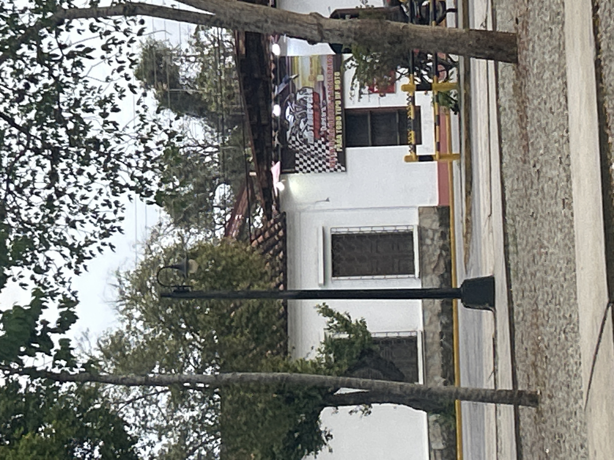 The image shows a white building with a tiled roof, likely a business, situated on a street in Santa Ana, Venezuela. The building features dark-colored windows with decorative metal grates. A prominent sign on the building displays a checkered flag and an image of motorcycles, with text that appears to be in Spanish, possibly indicating a motorcycle-related business such as sales or repair. The street in front of the building is paved with asphalt and has a gravelly shoulder. A black lamppost stands on the sidewalk. Trees with green foliage are visible in the foreground and background, suggesting an outdoor, daytime scene. A yellow and black striped barrier is positioned along the sidewalk, and a glimpse of parked motorcycles is visible in the background.