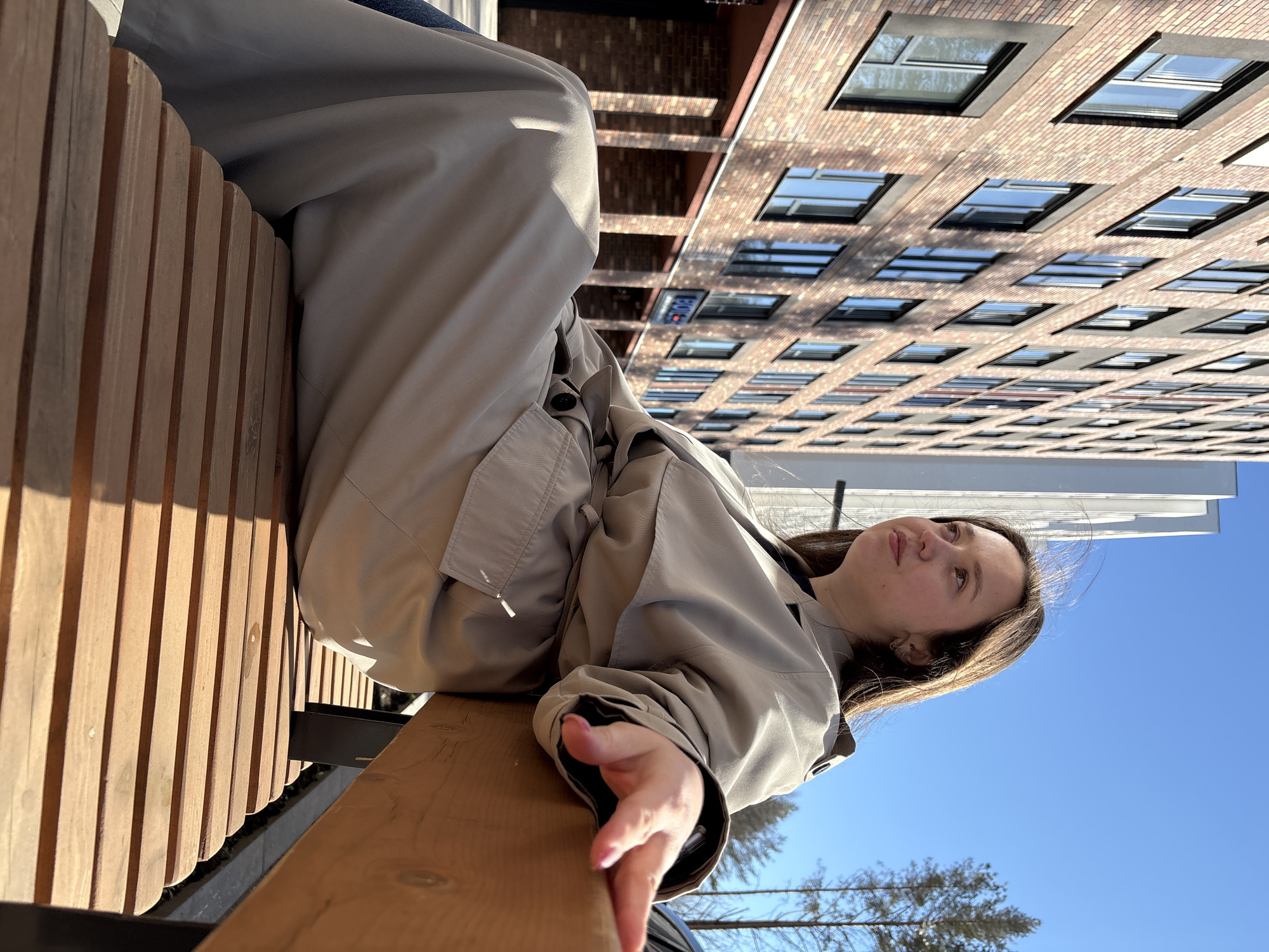 A woman sits on a wooden bench in Izhevsk, Russia, with a brick building in the background. The woman is wearing a beige trench coat and her dark hair is partly visible. Her hand is extended over the edge of the bench. The sky is blue with a few trees visible in the distance. The brick building has numerous windows. The bench appears to be made of vertical wooden slats.