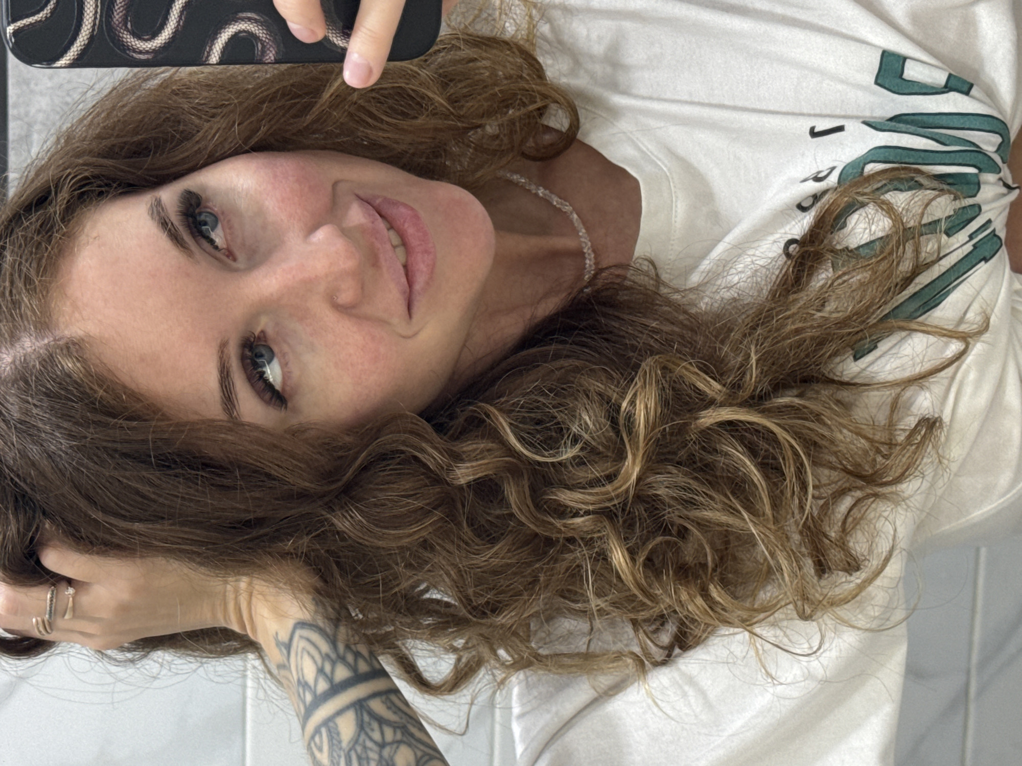 The image is a close-up, selfie-style photograph of a woman with long, curly brown hair. She is wearing a white t-shirt and a delicate beaded necklace. Her left hand is raised, touching her hair, and she is wearing a ring on her index finger. A tattoo is visible on her left arm. She is looking up and to her right, with a slight smile. A smartphone with a snake-patterned case is partially visible in the right side of the frame, held by her right hand. The background appears to be a bathroom with white tiled walls. There is no discernible connection to the city of Chaikovsky, Russia in the image.