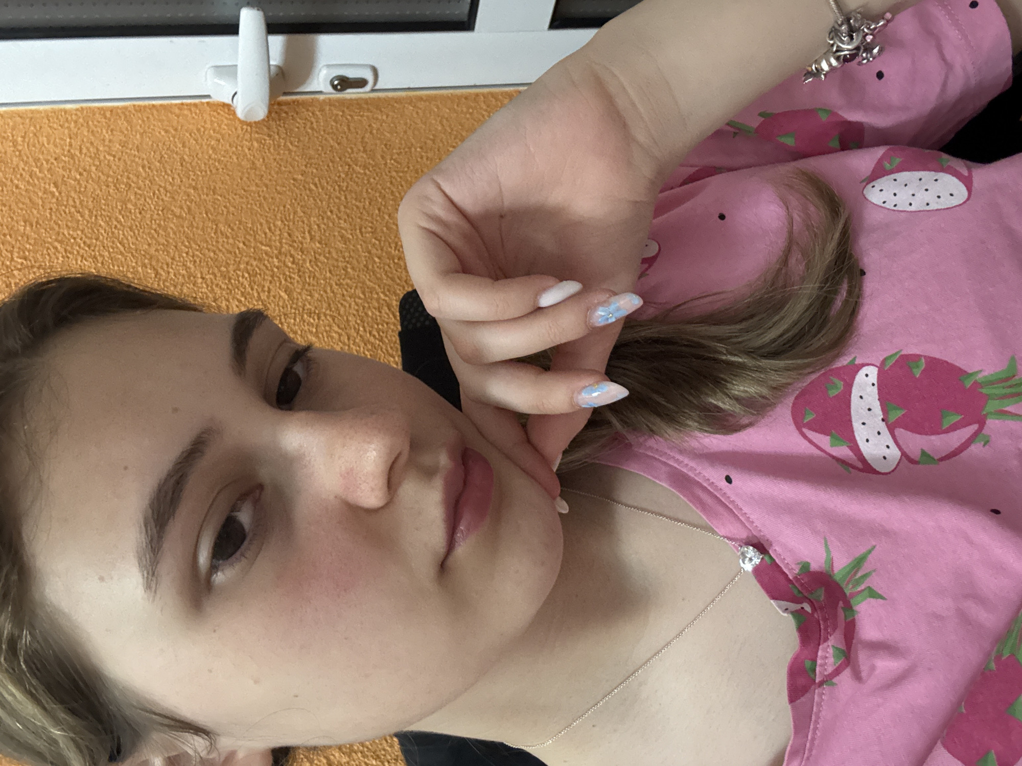 A young woman poses for a self-portrait photograph indoors. She has light skin, brown hair, and is wearing a pink shirt with dragon fruit patterns and a silver necklace with a diamond pendant. Her nails are decorated with blue floral designs. She is resting her face on her hand, looking at the camera. The background includes an orange wall, a white window frame, and a black object. The location is not identifiable as Panagyurishte, Bulgaria.