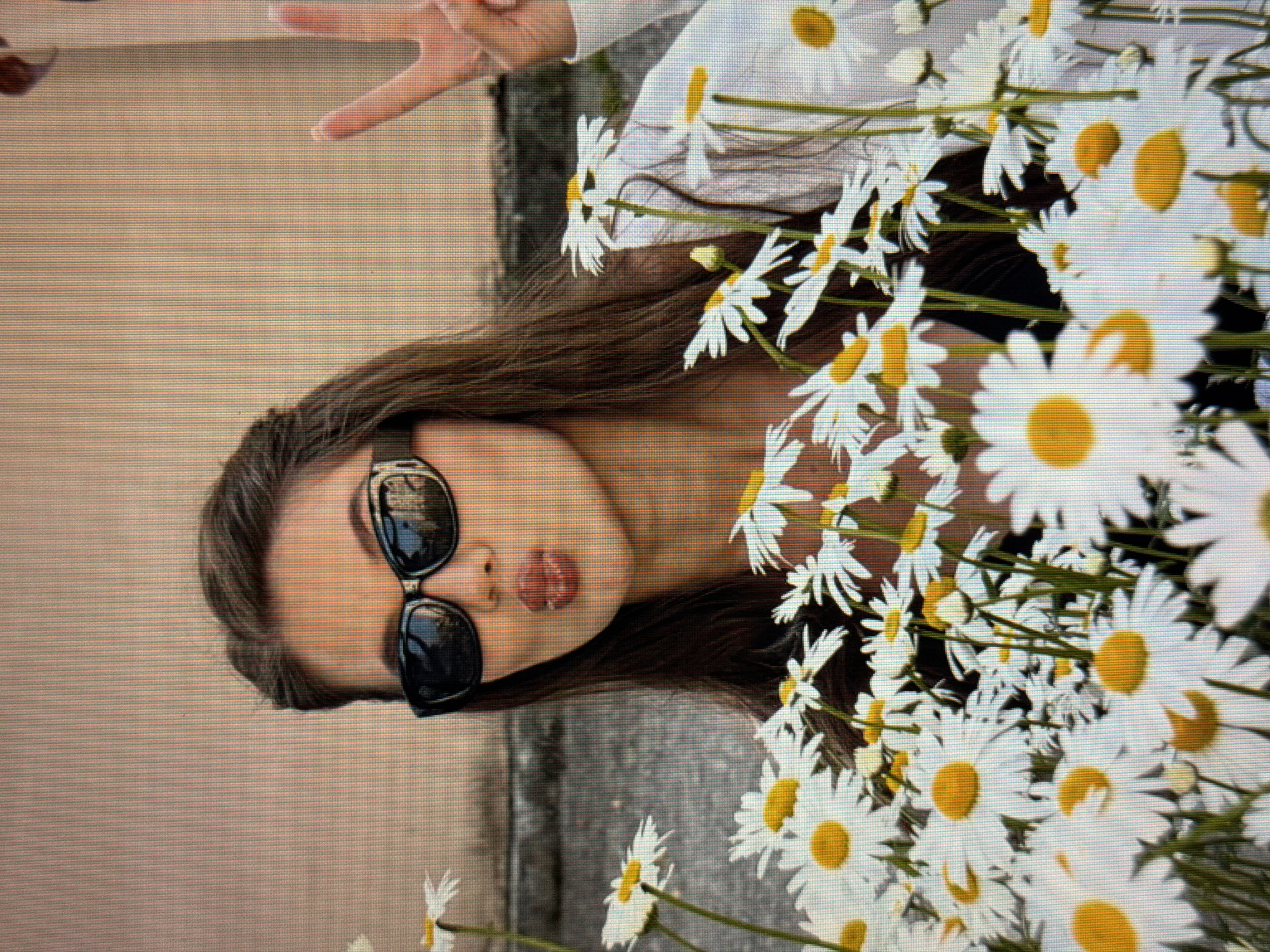 A woman with long, dark hair, wearing black sunglasses and a white shirt, poses behind a large bouquet of white daisies. She is puckering her lips and holding up two fingers in a peace sign. The background is a muted, light-colored wall. The scene is set outdoors.