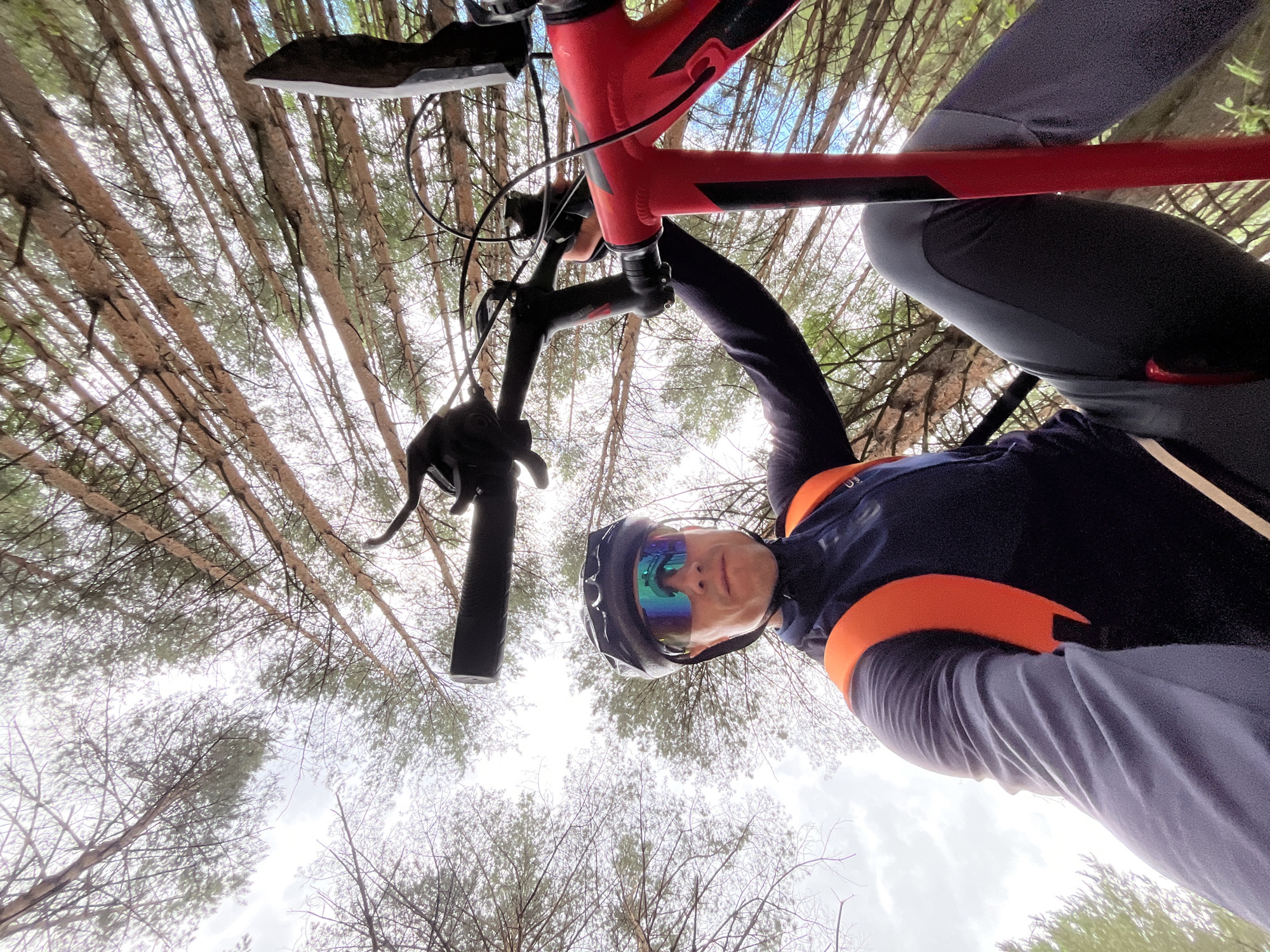 A low-angle, wide shot captures a male mountain biker in a forest environment, looking directly at the camera. The biker is wearing a black helmet, mirrored sunglasses with blue and green lenses, a dark blue long-sleeved jersey with orange accents on the shoulders and chest, and dark grey pants. He is seated on a red and black mountain bike, holding the handlebars. The background consists of a dense forest with tall, thin trees, their branches extending upwards and creating a canopy against a bright, overcast sky. The perspective suggests the photo was taken by the biker himself, using a wide-angle lens and holding the camera at arm's length. The scene is devoid of any city landmarks, indicating an off-road location.