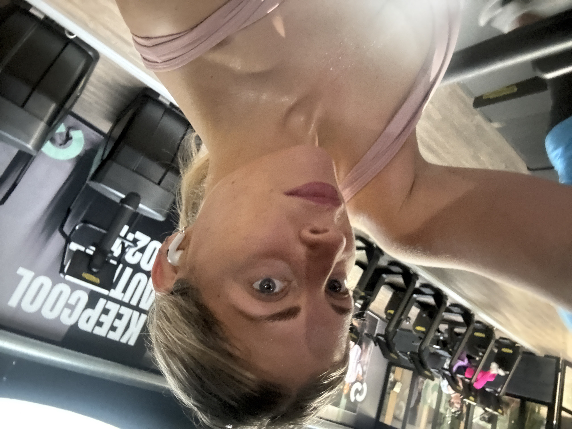A slightly out-of-focus, low-angle selfie of a Caucasian woman is taken in what appears to be a gym. The woman has fair skin, light-colored hair, and is wearing a light pink sports bra. Sweat is visible on her neck and chest. She is wearing a white wireless earbud in her left ear.
In the background, gym equipment is visible, including weight racks and treadmills. A sign with the words "KEEP COOL OUT 024" can be seen next to the weight racks. The overall setting suggests a modern, well-equipped gym. The location in Paris, France, cannot be determined from the visual information provided.