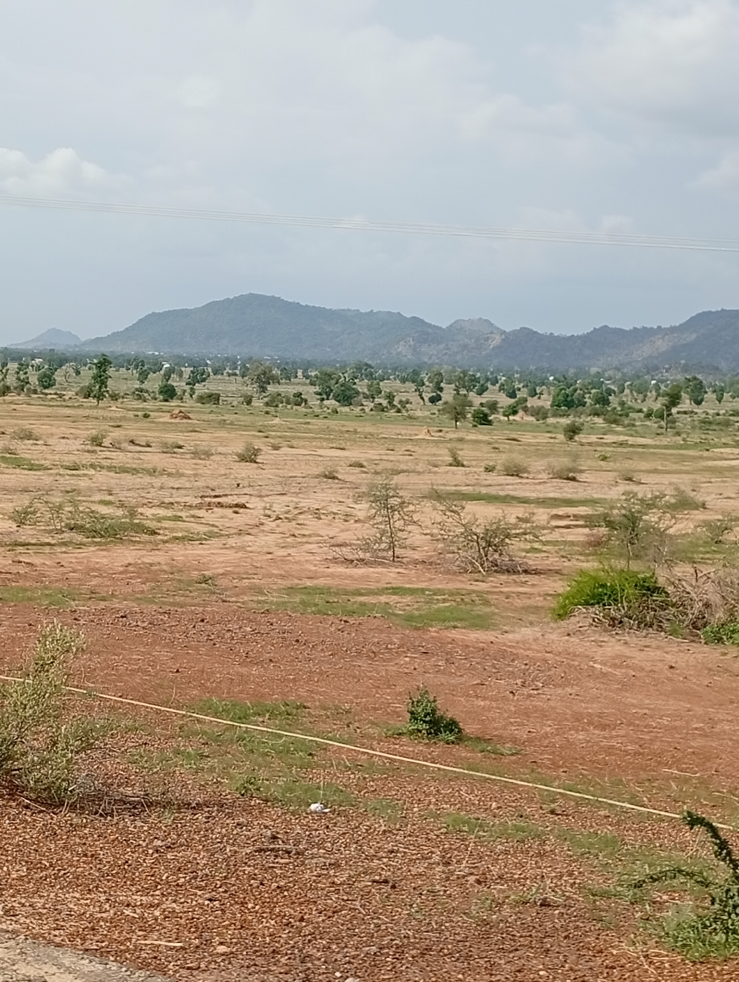 This is a landscape photograph taken in Shuwa, Nigeria. The image depicts a semi-arid environment with rolling hills in the background, sparsely populated by trees and vegetation. The foreground is characterized by dry, reddish-brown earth, interspersed with patches of dry grass and scattered shrubs. A thin, yellow line, possibly a rope or wire, runs horizontally across the mid-ground. In the far distance, a range of mountains is visible under a hazy, overcast sky with some clouds. The overall impression is of a rural or undeveloped area. There are no people or animals visible in the image.