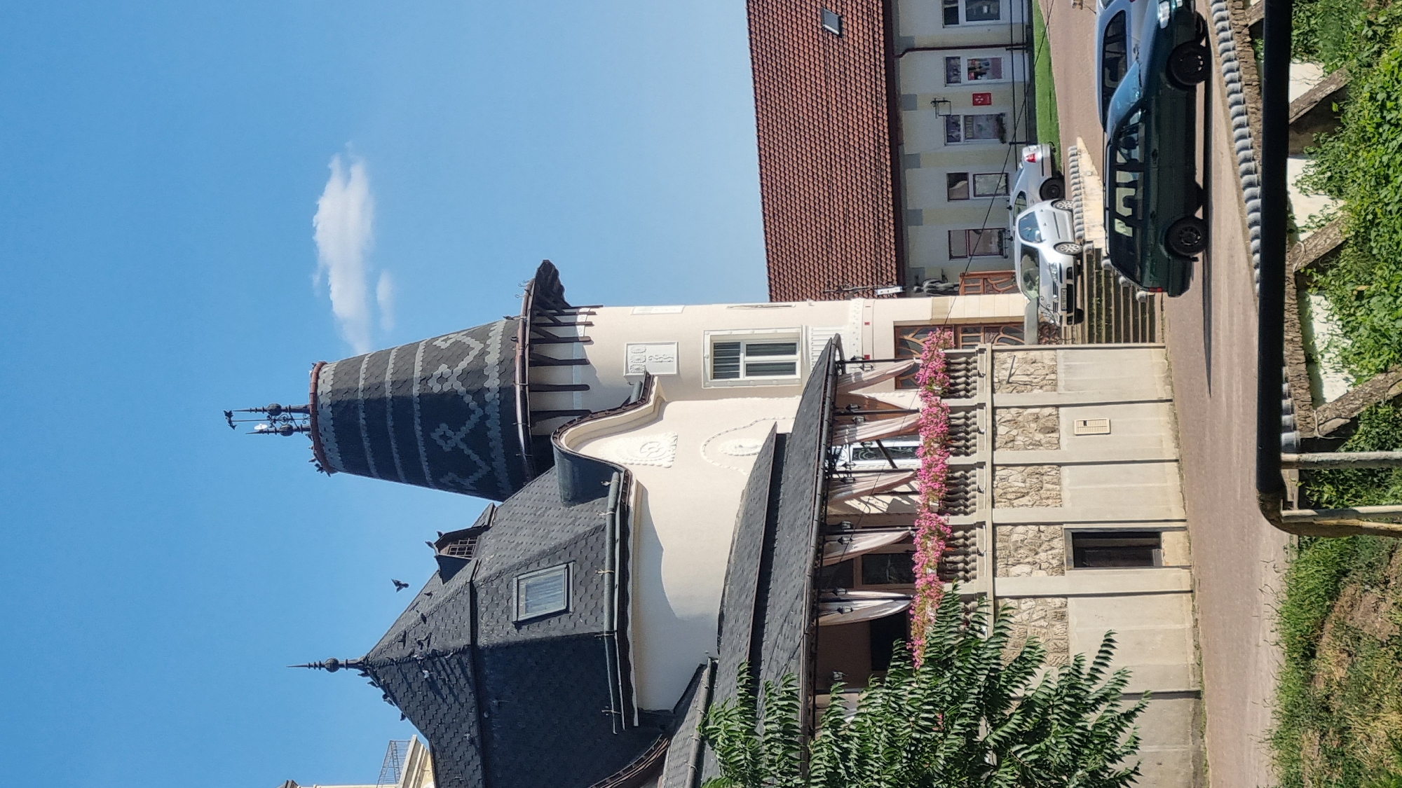 The image shows a large, ornate building with a dark, conical roof and a tall, pointed tower. The building has white walls and dark trim. There is a balcony with pink flowers and curtains. In front of the building, there are several parked cars and a set of stairs leading up to the entrance. A smaller building with a red tile roof is visible in the background. The sky is blue with a few white clouds. This building is identified as Hotel Vila San Remo in Berehove, Ukraine.