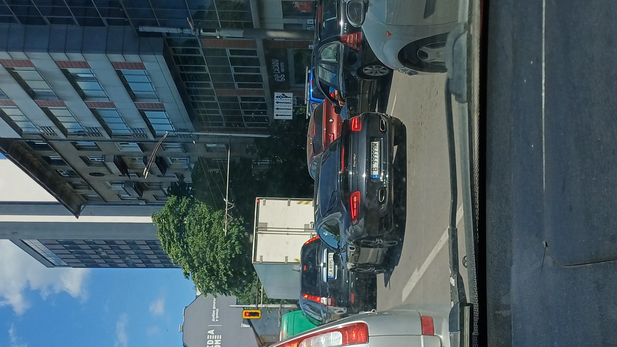 The image depicts a street scene in Sofia, Bulgaria, likely during daytime given the bright lighting. The perspective appears to be from inside a vehicle, looking forward and slightly to the right. The foreground is dominated by the dark interior of a car, with part of its dashboard and windshield visible.
The main focus of the scene is a traffic jam on a paved road. Several cars are lined up, indicating a standstill. Among the visible vehicles is a black Bentley, its license plate reading "B 99999M". Next to it is a red car, and further down the line are other vehicles, including a white truck and a black car with license plate "CA 4895 TT". In the background, modern buildings with glass facades and windows are visible, suggesting an urban environment. One building has a sign with the text "CALADU". Trees with green foliage are also present, adding a touch of nature to the urban landscape.
A traffic light, showing yellow, is visible on the left side of the road, indicating potential changes in traffic flow. The sky is partly cloudy, with patches of blue. The overall impression is a typical city traffic situation on a bright, presumably warm day. There are no people prominently visible, but the presence of multiple cars implies their existence within the scene.