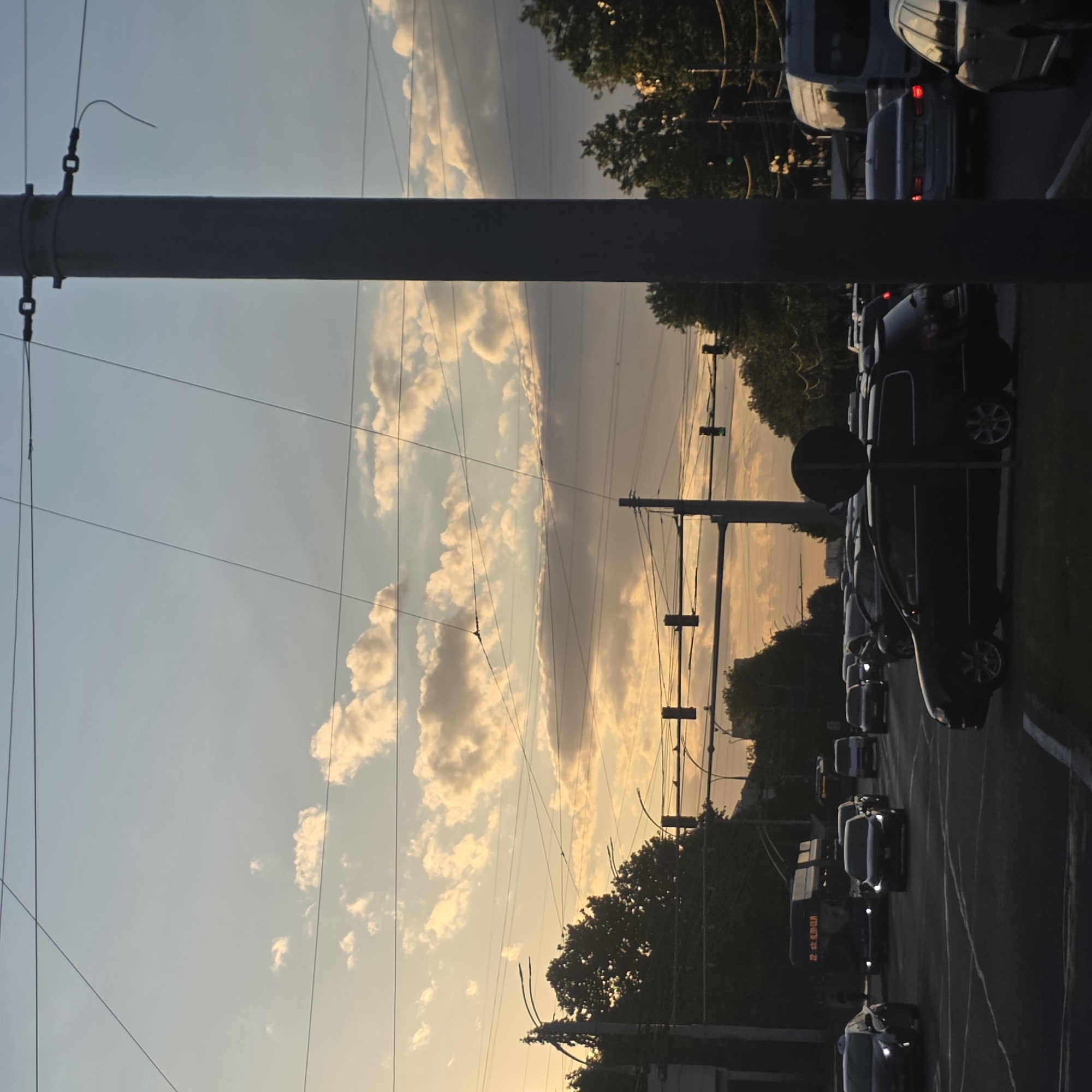 The image depicts a street scene at dusk in a city, possibly Unknown, Unknown. A large utility pole stands prominently in the foreground to the right, with overhead wires crisscrossing the sky. The sky is filled with scattered clouds, illuminated by the warm tones of the setting sun, creating a blend of pale blue and orange hues.
The street is busy with traffic. Several cars are visible, including a dark-colored station wagon in the lower half of the frame, and a light-colored sedan further back. A trolleybus with the number "22" and Cyrillic text is seen in the middle ground. Traffic lights are present, with one showing a green light on the right side of the frame. Trees line the sides of the road, and buildings are faintly visible in the distance. The overall impression is one of a typical urban environment during the evening rush hour.
