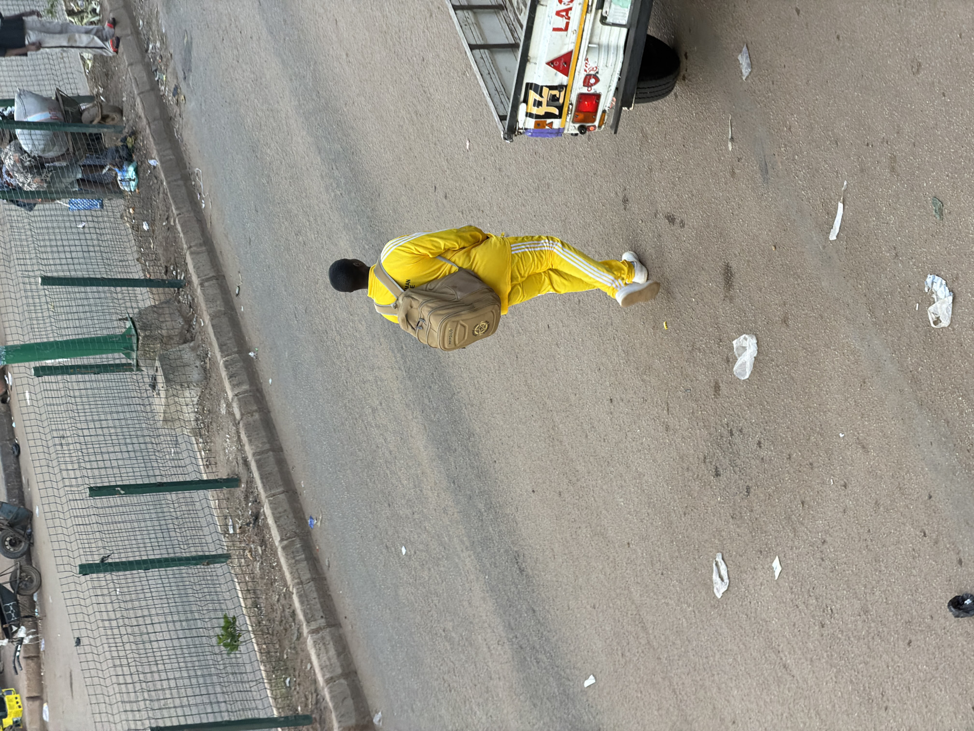 A person is seen wearing a bright yellow tracksuit and white sneakers. They are carrying a beige backpack. The person is standing on a paved road, which appears to be a street or a roadside. The setting is outdoors, with a truck visible in the background. The truck has text on its side, but it is partially obscured and difficult to read. The road is somewhat littered with small pieces of paper or debris. A metal fence is also present along the side of the road, with some vegetation growing behind it. The lighting suggests it is daytime. The overall scene appears to be a candid street photograph, possibly in an urban or semi-urban environment in Bukuru, Nigeria.