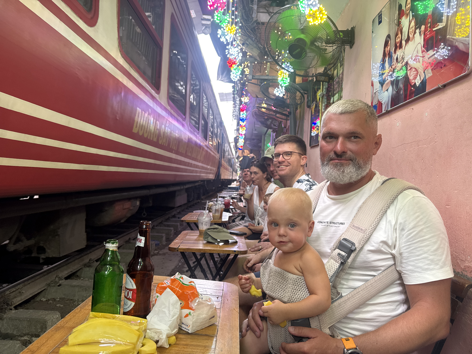 This image captures a vibrant scene at a trackside cafe on Hà Nội's famed Train Street, Vietnam. In the foreground, a light-skinned adult male with a short grey beard and close-cropped grey hair looks directly at the camera. He wears a white t-shirt with "CONCRETE STRUCTURES" and "HEART OF FUNCTIONALITY" printed on it, and an orange-strapped wristwatch on his left wrist. He is supporting a beige BabyBjörn baby carrier, which holds a fair-skinned infant with blonde hair and blue eyes. The infant, wearing a light-colored mesh top, is also looking at the camera while holding and eating pieces of yellow fruit.
On a small, low wooden table directly in front of the man and baby are a green glass bottle and a brown glass bottle, both appearing to contain beer. Also on the table are several slices of plastic-wrapped yellow fruit, an orange snack bag, and some crumpled white tissue paper.
To the left, a large red and white passenger train, bearing the gold Vietnamese text "DUONG SAT VIET" (VIETNAMESE RAILWAY), is positioned on the tracks, running parallel and extremely close to the cafe's seating area. The train's windows are visible. The train tracks themselves are evident beneath the tables and extending into the background.
Behind the main subjects, several other individuals are seated at similar small wooden tables, extending down the length of the trackside. One man with glasses and a dark shirt smiles towards the right, and a woman is seated behind him.
The environment is characterized by a narrow passage. Above the cafe seating, numerous strings of colorful decorative LED lights in various shapes and hues (including pink, green, yellow, white, and blue) are strung overhead. On the right-hand wall, which is painted a light pink or peach color, two green oscillating fans are mounted. A large, reflective framed picture featuring several women is also affixed to this wall. The ceiling appears to be a simple, perhaps corrugated, structure with visible wiring.
