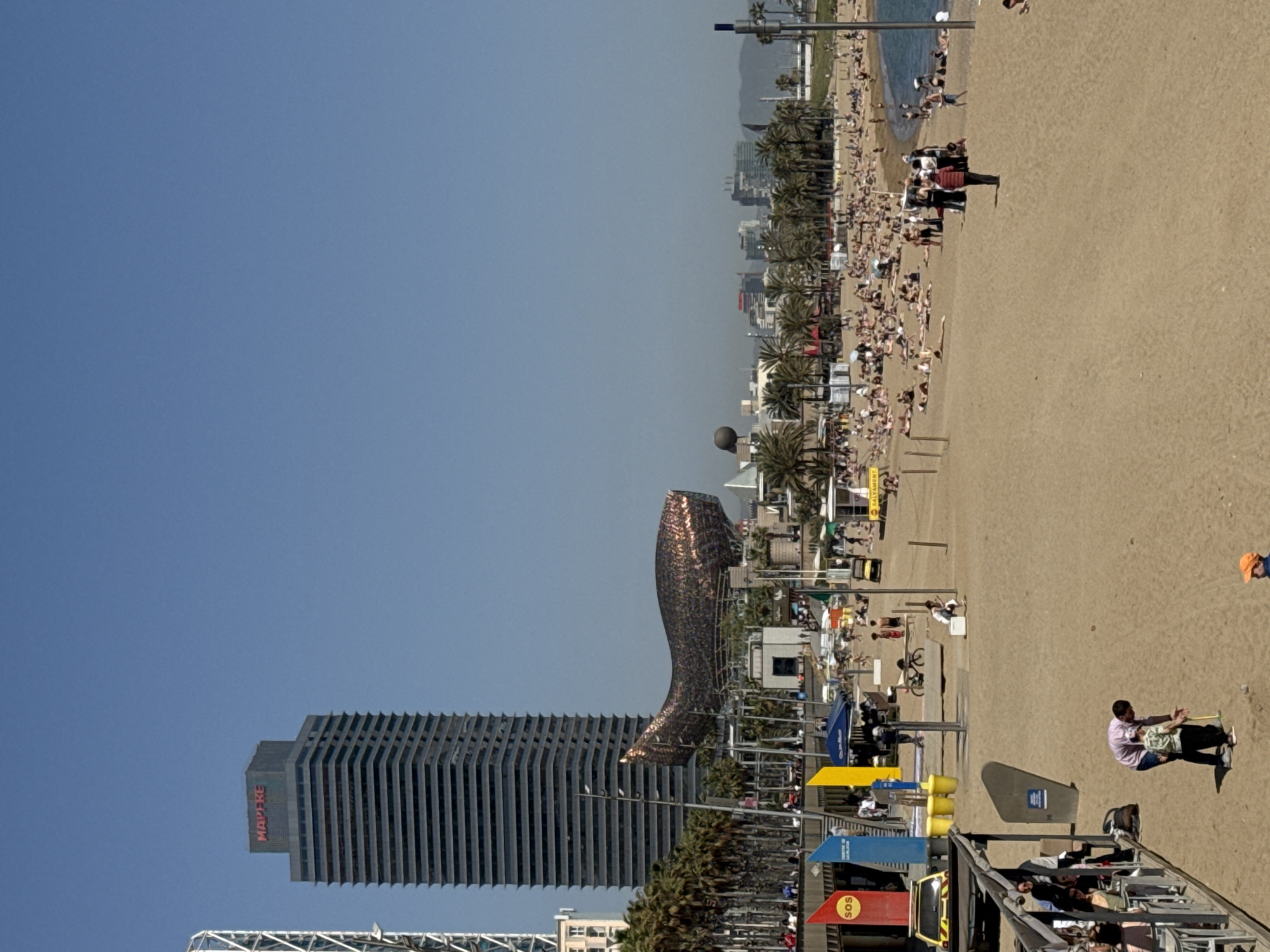 The image shows a view of Barceloneta Beach in Barcelona, Spain, on a sunny day. In the foreground is a beach crowded with people. Further back, the distinctive Golden Fish sculpture is visible next to the Hotel Arts skyscraper. The sky is clear and blue.