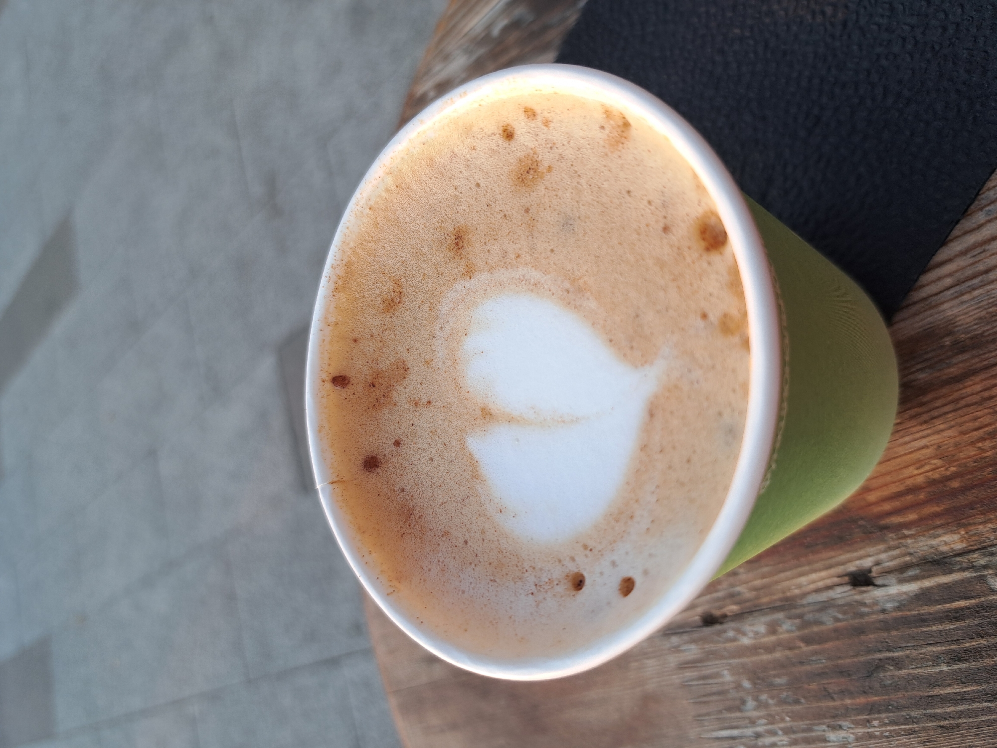 A close-up, eye-level shot shows a disposable paper cup with a green exterior and a white rim. The cup is filled with a frothy, light brown beverage, likely coffee. A heart shape is delicately etched into the foam, a common latte art design. The surface of the drink displays small bubbles and darker specks, possibly coffee grounds or cinnamon. The cup rests on a rustic wooden surface, its grain visible and worn. To the right of the cup, a dark, textured placemat or coaster is partially visible. In the background, a paved surface with rectangular stone tiles suggests an outdoor setting, possibly a sidewalk or patio in a city. The lighting indicates it might be late afternoon or early morning, with warm tones highlighting the scene. The image's composition and elements do not provide any specific information to pinpoint its location within Irkutsk, Russia, or any other city.