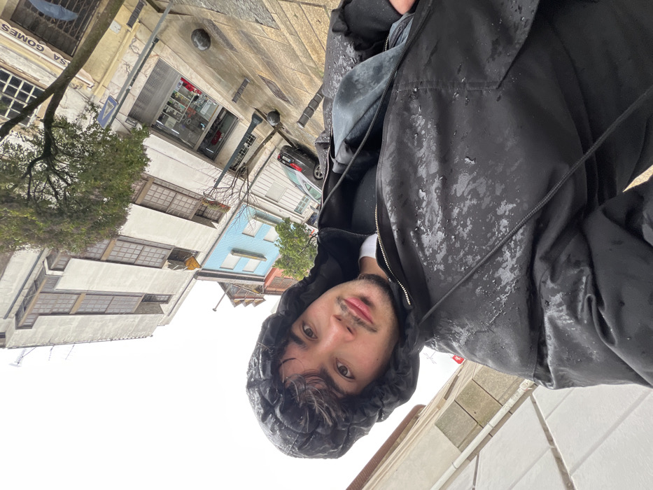The image captures a selfie of a male individual on a city street in Bragança, Portugal, under overcast, rainy conditions. The subject, a male estimated to be in his late teens or early twenties, has dark curly hair and light facial hair, including a mustache and goatee. He is wearing a black hooded jacket, with the hood pulled over his head; visible water droplets and streaking on the jacket surface indicate exposure to rain. He looks directly at the camera with a neutral expression.
In the background, a wet, textured paved street or sidewalk is visible to the right, showing reflections. Two dark, spherical bollards are positioned on the pavement, and a black hatchback vehicle is parked along the street. The street is lined with multi-story buildings: a prominent light-colored building with numerous rectangular windows dominates the upper background, exhibiting signs of wear. Adjacent to it on the right is a smaller, light-blue two-story building, followed by a partially visible building with an orange-colored facade. A ground-floor shopfront with a glass display and a sign reading "CASA GOMES" is visible. A tree with some green foliage and bare branches stands between the buildings and the street. The overall environment is urban, characterized by the wet surfaces and architectural details consistent with a European city street.