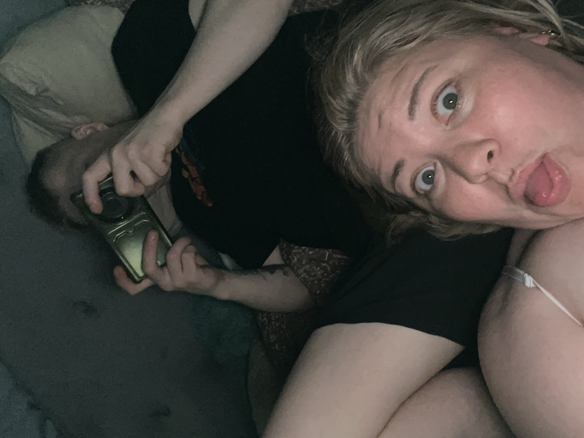 This indoor selfie features two individuals in a relaxed, low-light setting, likely a home, with a tilted perspective. In the foreground, on the right, a woman with light-colored hair is visible from the chest up, making a playful or surprised expression with wide eyes and her tongue slightly out. She is wearing a light-colored strap garment. Next to her, on the left, a man is partially visible, lying down. He has dark, short hair and is looking downwards, possibly at a gold-colored smartphone he is holding with an extended arm, which reveals a tattoo. A light-colored pillow is visible behind his head. The dim lighting suggests the photo was taken in the evening or at night. The scene captures a casual, candid moment between the two people. No text is visible in the image.