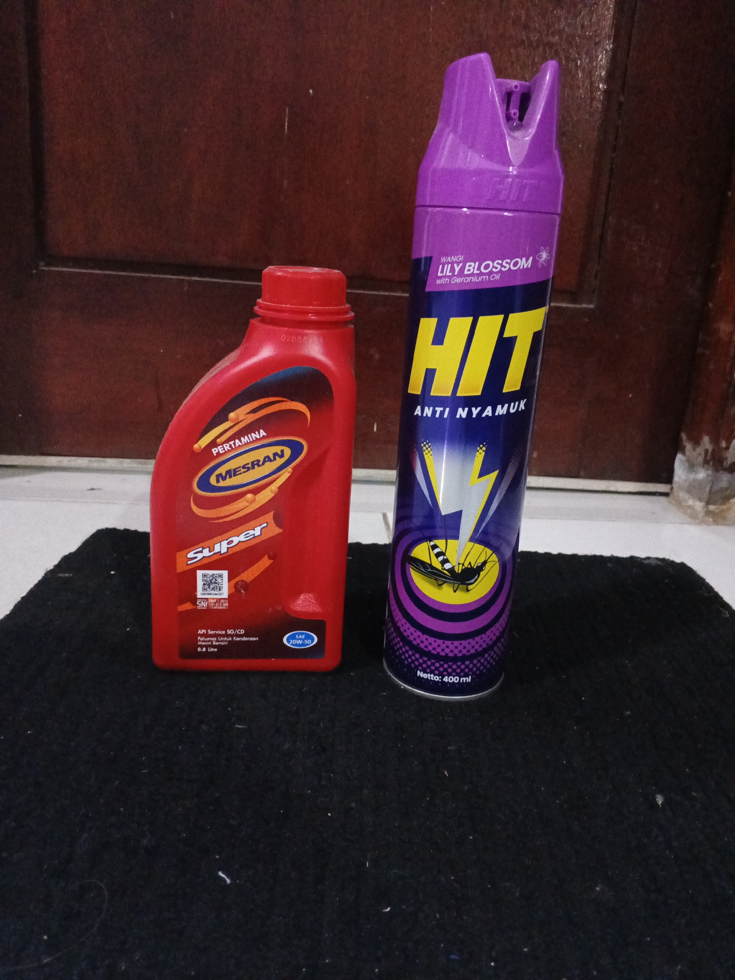 In Kemangsen, Indonesia, a still life image captures two distinct items placed on a dark, textured surface. To the left, a bright red bottle with a white label features the brand name "MESRAN" in blue oval, with "PERTAMINA" above and "Super" in bold orange lettering below. The bottle appears to be motor oil, indicated by text like "API Service SG/CD" and "Pelumas Untuk Kendaraan Mesin Bensin". A QR code is also visible on the label. To the right, a tall, purple aerosol can stands upright. The can has a purple cap and the prominent yellow lettering "HIT" against a dark blue background. Text above "HIT" reads "WANGI LILY BLOSSOM with Geranium Oil", and below it says "ANTI NYAMUK". The can also displays a graphic of a mosquito being zapped by lightning and has the text "Netto: 400 ml" at its base. The background consists of a dark brown, wooden door with a subtle grain.