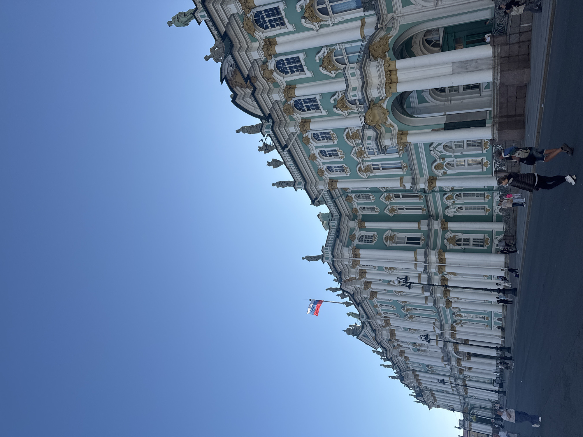 This media file captures a low-angle view of a grand, ornate building with a light teal facade, white columns, and gold accents. The architectural style suggests a historical European palace or museum. Multiple windows with decorative frames are visible along the facade, and statues or sculptures adorn the roofline. A Russian flag is prominently displayed on a flagpole.
Several people are present, appearing as small figures against the scale of the building. They are walking on a paved area in front of the structure, suggesting it is a public space, possibly a square or a street adjacent to the building. The people are dressed casually in t-shirts, shorts, and pants, indicating warm weather. One person is walking with a backpack.
The scene is illuminated by clear, bright sunlight, suggesting it is daytime, likely morning or afternoon, with a cloudless, deep blue sky dominating the upper portion of the frame. The shadows cast by the building and the lampposts indicate the sun is relatively high.
The overall mood is bright and serene, with the magnificent architecture and clear sky creating a sense of grandeur and pleasantness. The presence of people suggests it's an active public area. The context clues, such as the Russian flag and the architectural style, point to a location in Saint Petersburg, Russia, likely a significant landmark like the Winter Palace. No visible text is present in the media file.