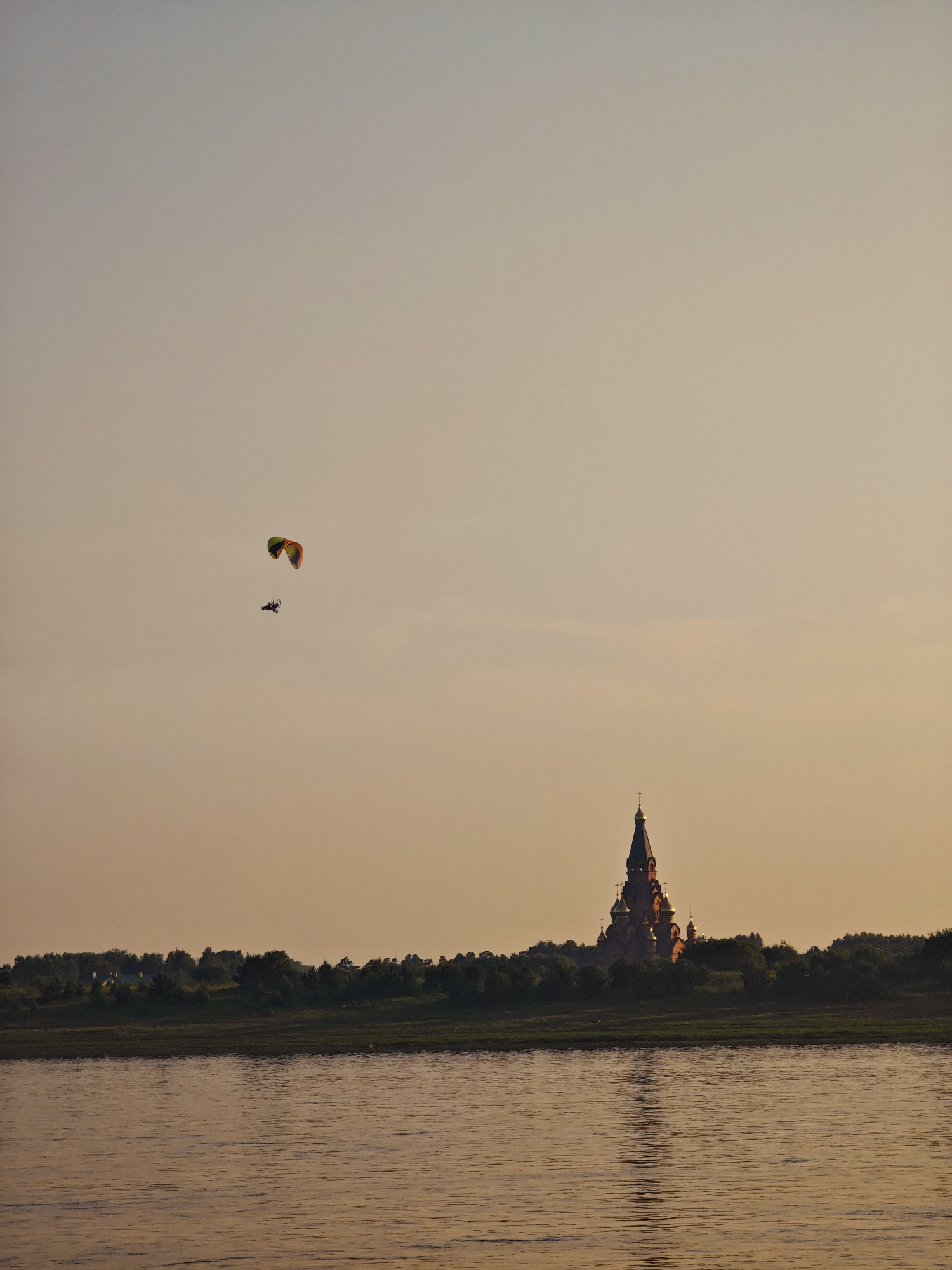 The image shows a paraglider in flight against a pale orange sky, with a silhouette of a large, domed building, likely a church, visible on a tree-lined horizon. The paraglider, with its colorful wing, is positioned in the upper left quadrant of the frame. Below the paraglider and to the right, the church structure is illuminated by a warm light, casting a reflection on the rippling water in the foreground. The water takes up the bottom third of the image, and its surface reflects the soft hues of the sky and the distant land. The scene appears to be a serene landscape at dusk or dawn, possibly along a river. This building, the Church of the Assumption of the Blessed Virgin Mary, is a prominent landmark in Lesosibirsk, Russia.