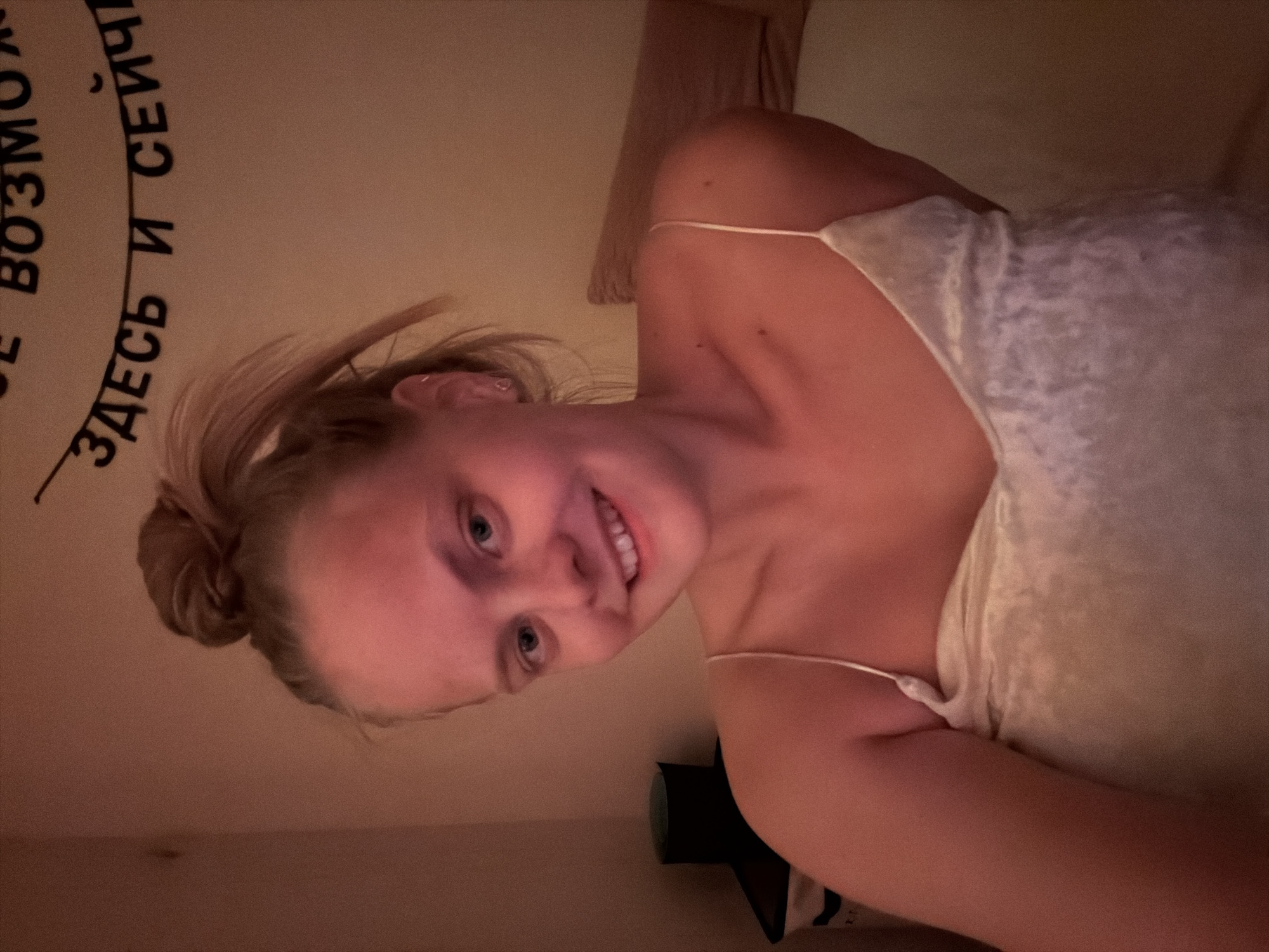 A young woman is shown in a medium, indoor shot, appearing to lie in bed, likely in a private residence. She has blonde hair pulled up in a bun and is smiling at the camera. She is wearing a light-colored camisole. In the background, some Cyrillic text is visible on the wall, and there is a bedside object on a surface next to her. There is no verifiable information in the image to confirm a location in Izhevsk, Russia.