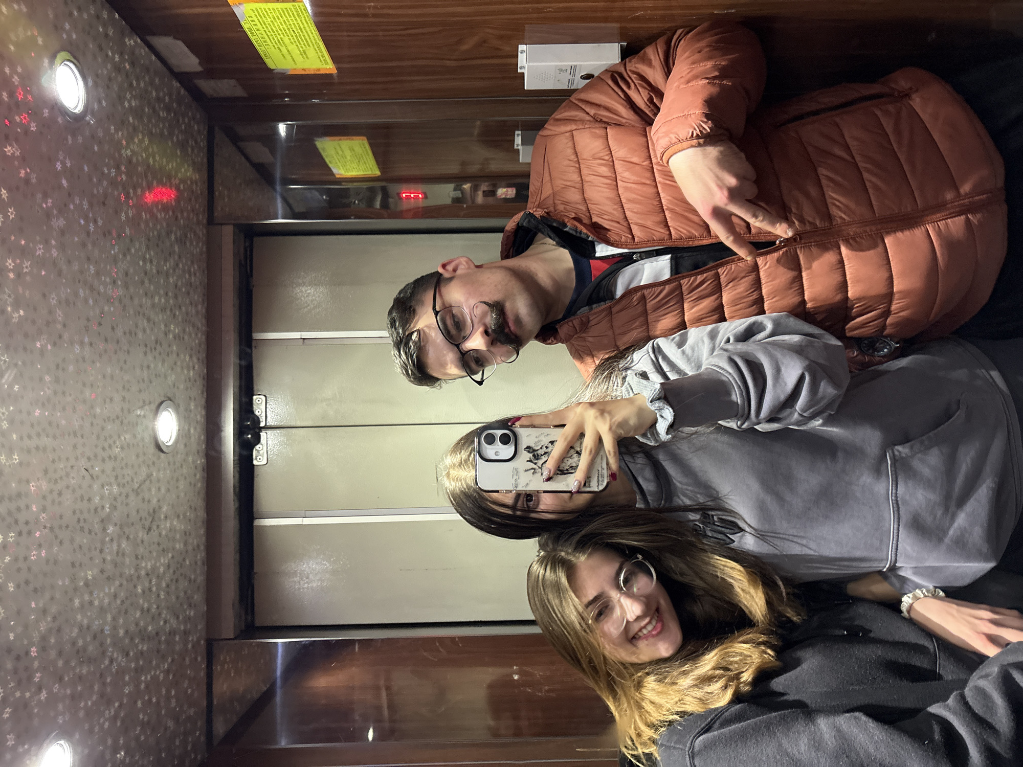 Three individuals are taking a selfie in what appears to be an elevator. The location, Blagoevgrad, Bulgaria, cannot be determined from the visual information provided. A man in a brown jacket with glasses poses at the top of the frame. A woman holds a smartphone with a white phone case to take a selfie and another woman in glasses is positioned at the bottom of the frame. The elevator has mirrored walls, revealing the elevator doors in the background and lighting features on the wall.