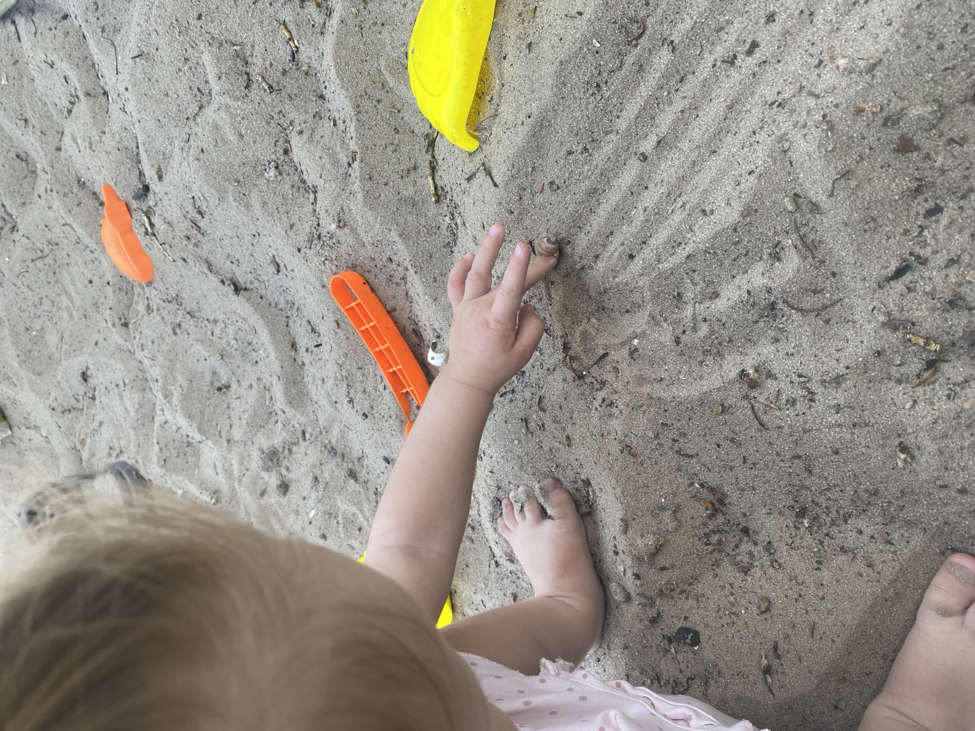 The image shows a toddler playing in the sand. The toddler's head and hair are visible in the upper left corner of the frame, with their right arm reaching out towards the sand. Their feet are also visible in the lower left and bottom of the frame, with sand on their toes. In the sand, there are several brightly colored plastic toys, including an orange shovel and a yellow scoop. The sand itself appears to be damp and mixed with small debris like twigs and small stones. The scene suggests a playful outdoor activity, possibly in a sandbox or a sandy area. The image does not provide any specific visual cues to determine the location within the city of Dnipro, Ukraine.