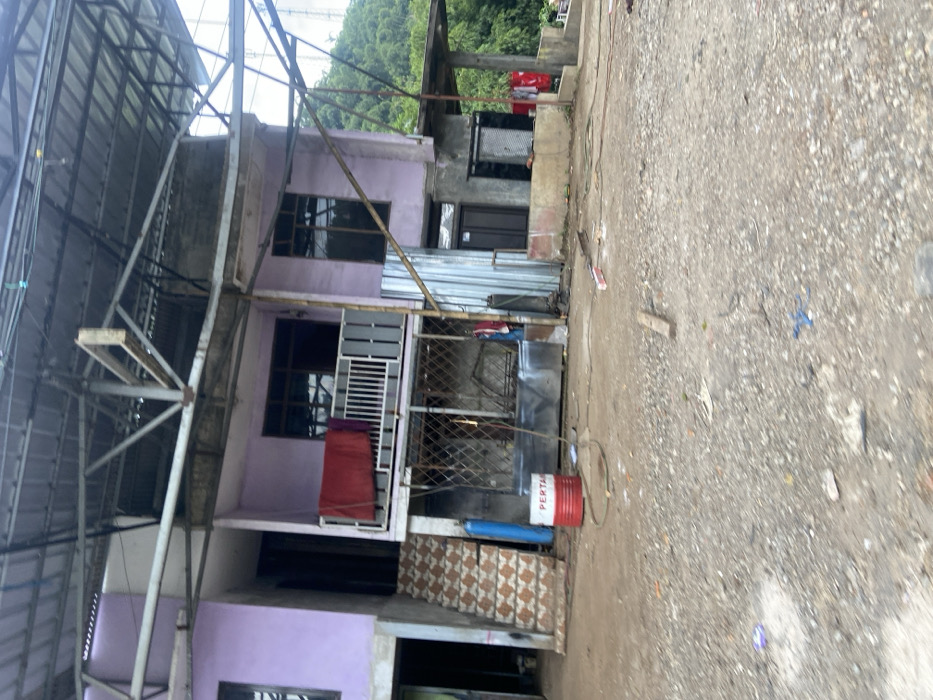 The image shows a two-story building with a pink exterior and a metal roof. The building is located in Cipatat, Indonesia, at coordinates -6.792 latitude and 107.434 longitude. The building is partially obscured by a large metal frame structure. The building has a balcony with a white railing, a set of stairs leading up to the balcony, and a window with a red curtain on the second floor. The ground below is covered in gravel and there is a red and white barrel with the word "PERTA" visible. In front of the building, there are some loose metal pipes and a few pieces of debris. There is a green hillside in the background. There are no people visible in the image.